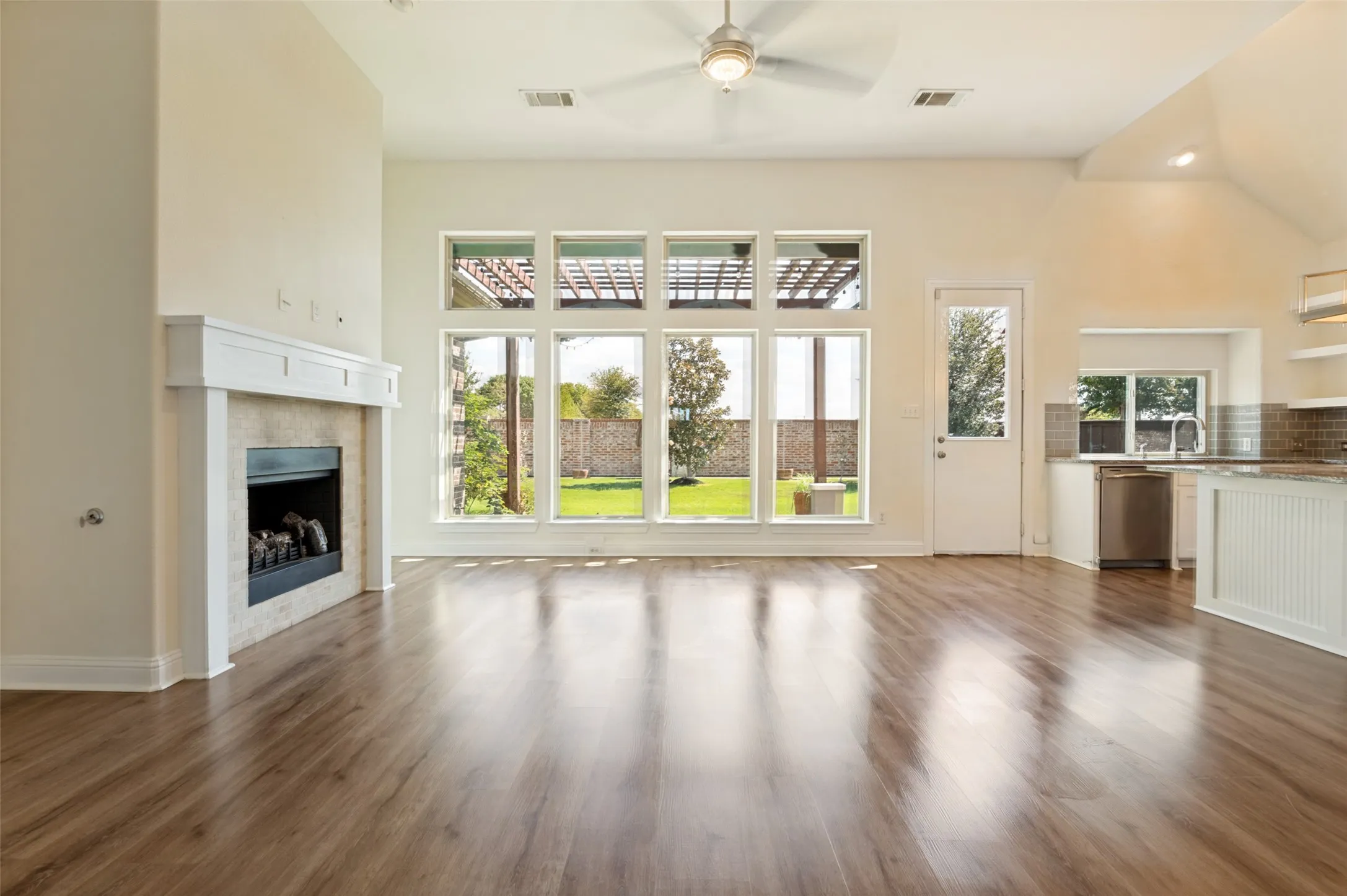 Vaulted ceiling with a ceiling fan, wood-finish flooring, fireplace with a white mantel, kitchen with a stainless steel dishwasher, and large windows overlooking the backyard
