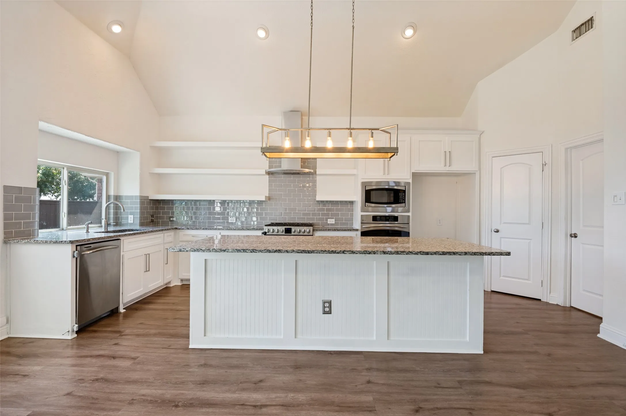 Chef's kitchen featuring white cabinetry with vertical beadboard detailing, granite countertops, a large center island with seating space, stainless steel appliances including double wall ovens, and a linear pendant light fixture