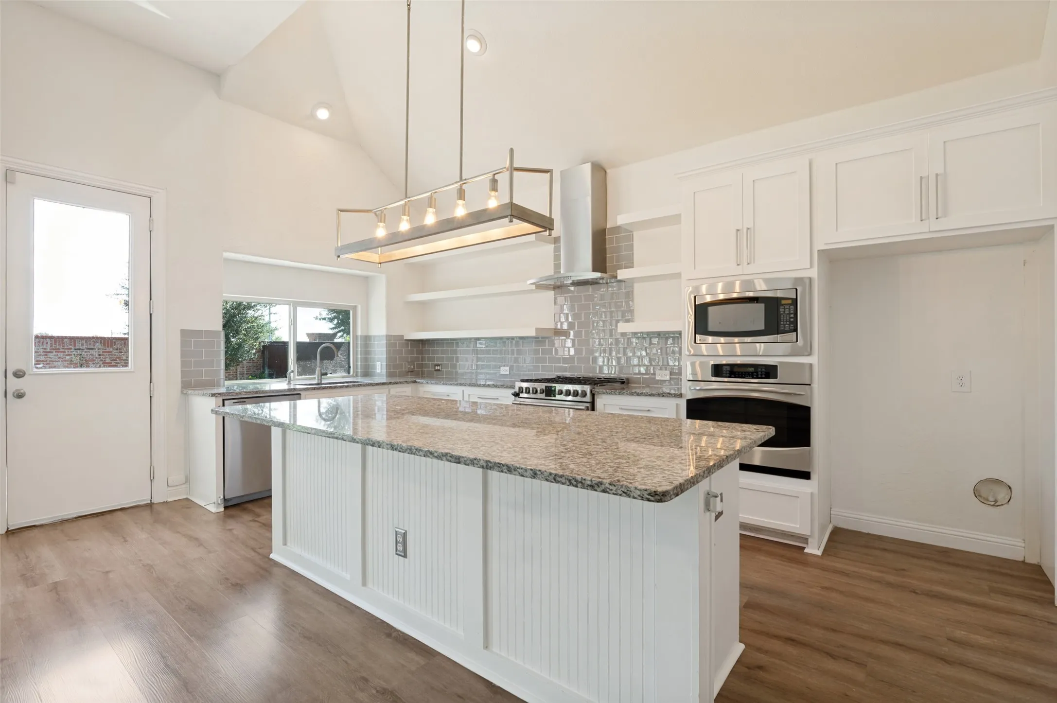 Vaulted ceilings with recessed lighting, wood-finish flooring, white shaker cabinetry, an eat-at kitchen island with a granite countertop, and stainless steel appliances