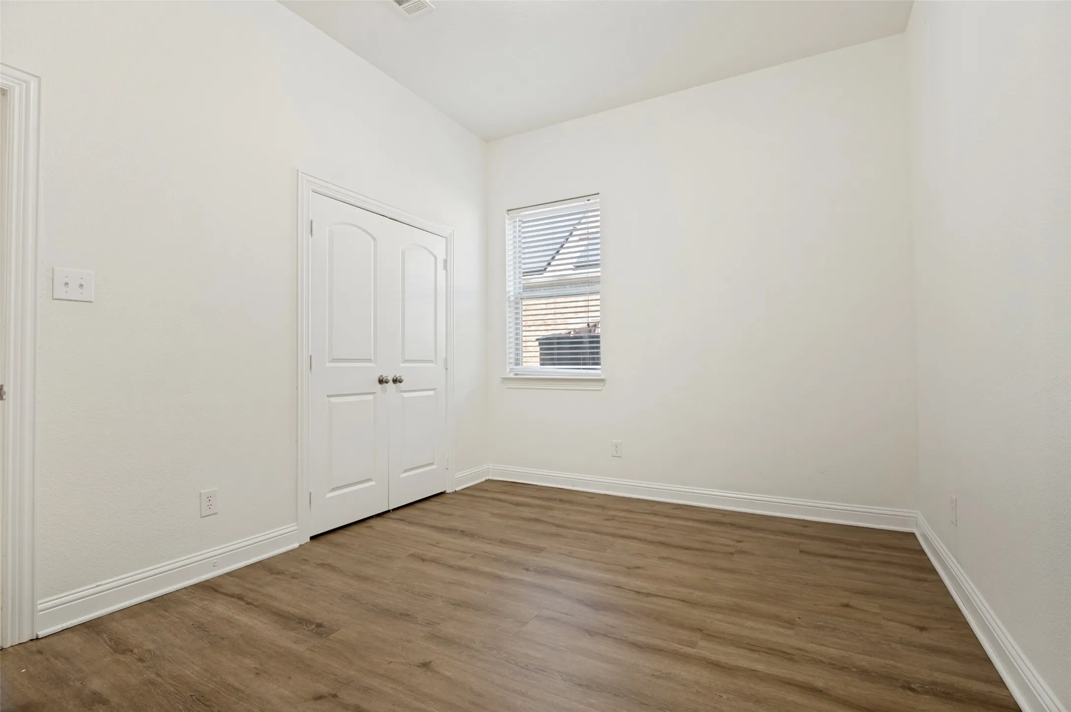 Bright, light-filled bedroom showcasing wood-finish flooring, white painted double-door closet with panel detailing, and a large window with operable blinds allowing abundant natural light