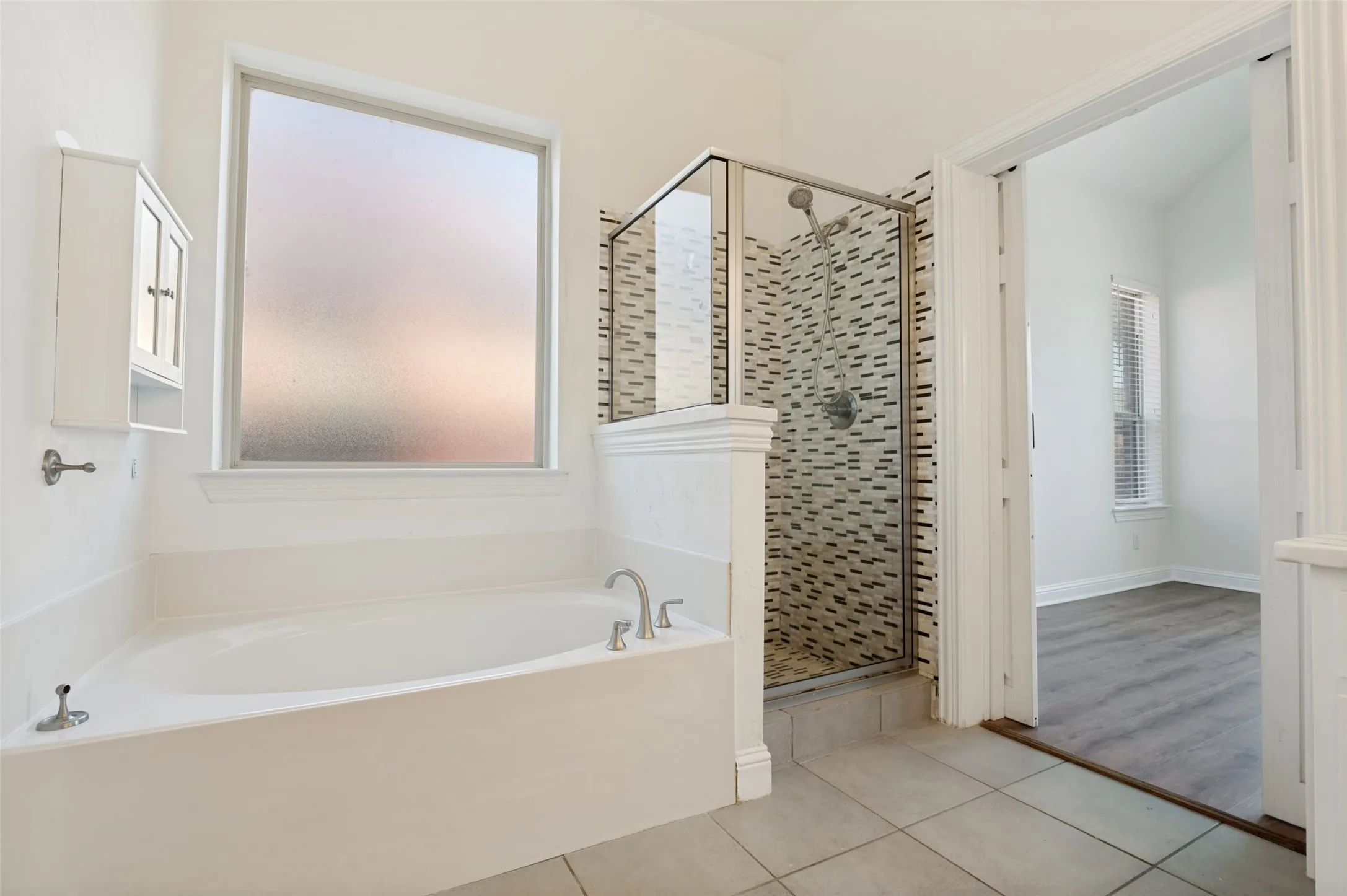 Built-in soaking tub with brushed nickel fixtures, separate glass-enclosed shower featuring mosaic tile, frost-glass window, tiled flooring, and wall-mounted storage cabinet