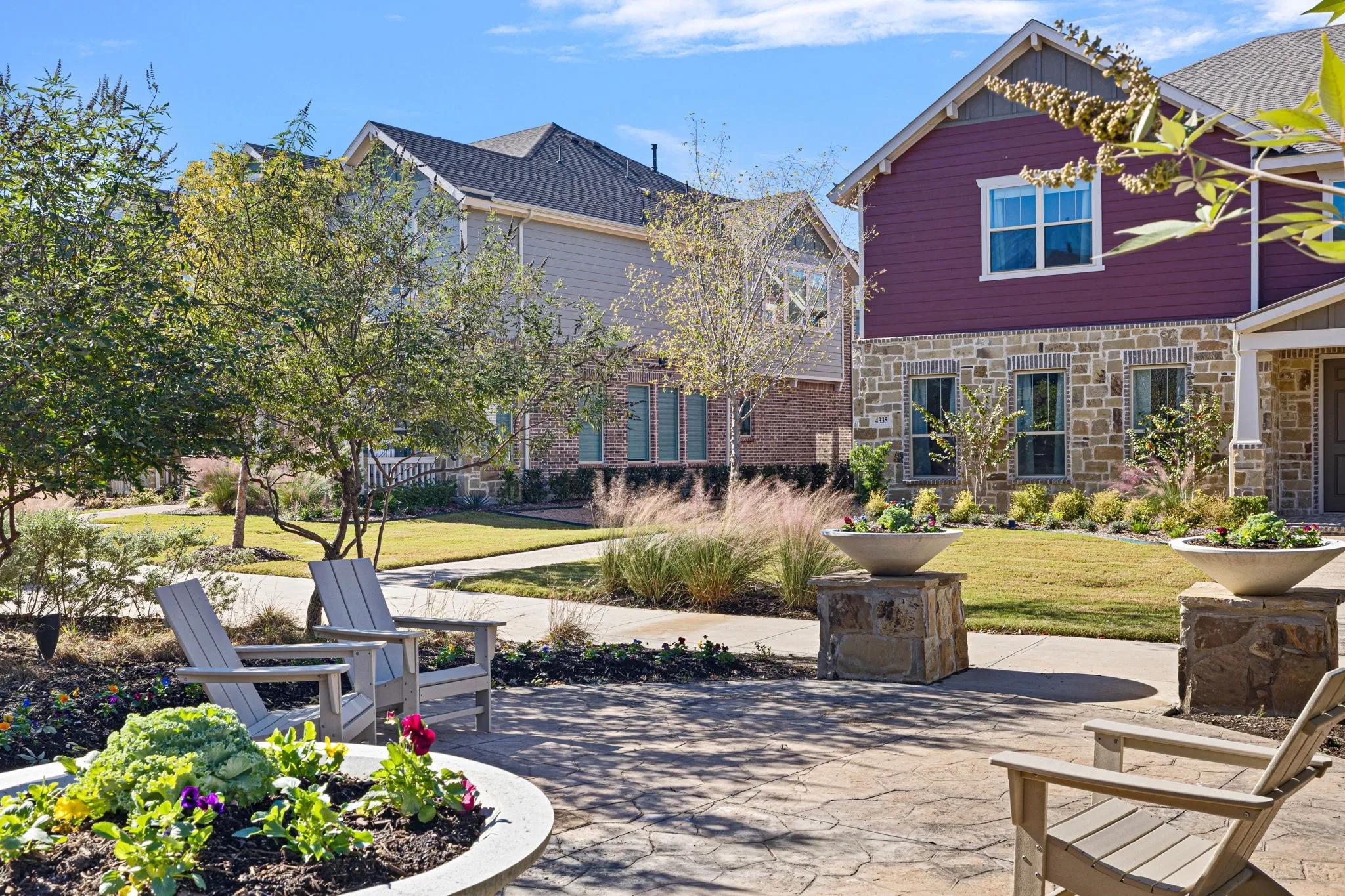 Paved patio area with a circular raised planter, mature trees, manicured lawn, and stone-clad planter pedestals