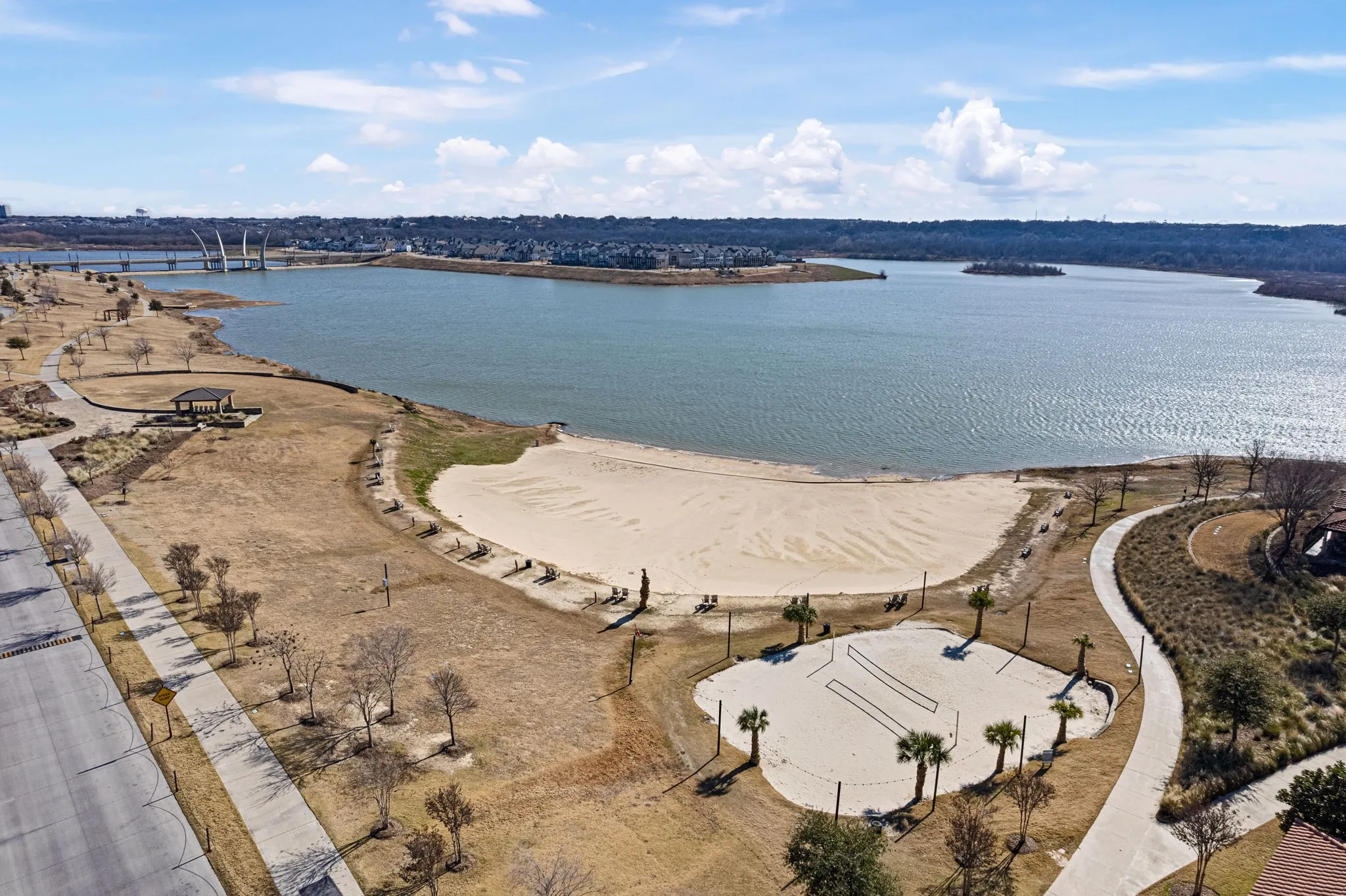 Lakeside recreational area featuring a sandy beach, sand volleyball court, pavilion, and paved walking paths