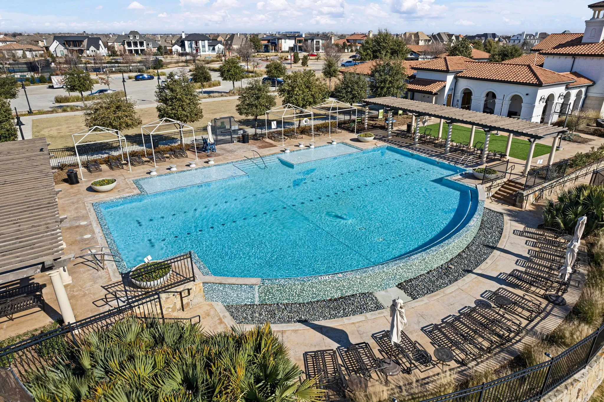Large swimming pool with integrated lap lanes, extensive paved pool deck, shaded pergola structure, and adjacent building with arched openings and a red tile roof