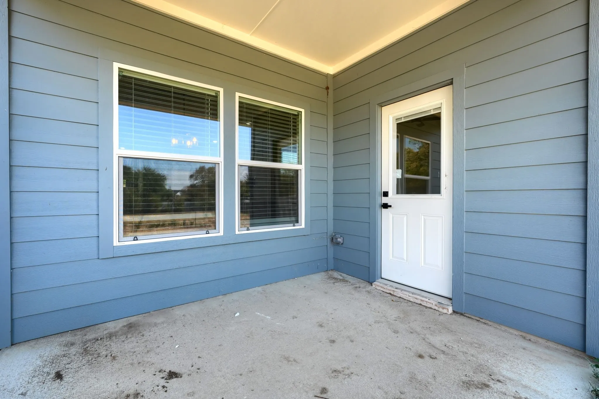 Covered patio featuring blue siding, two windows with blinds, a white exterior door, and a concrete surface