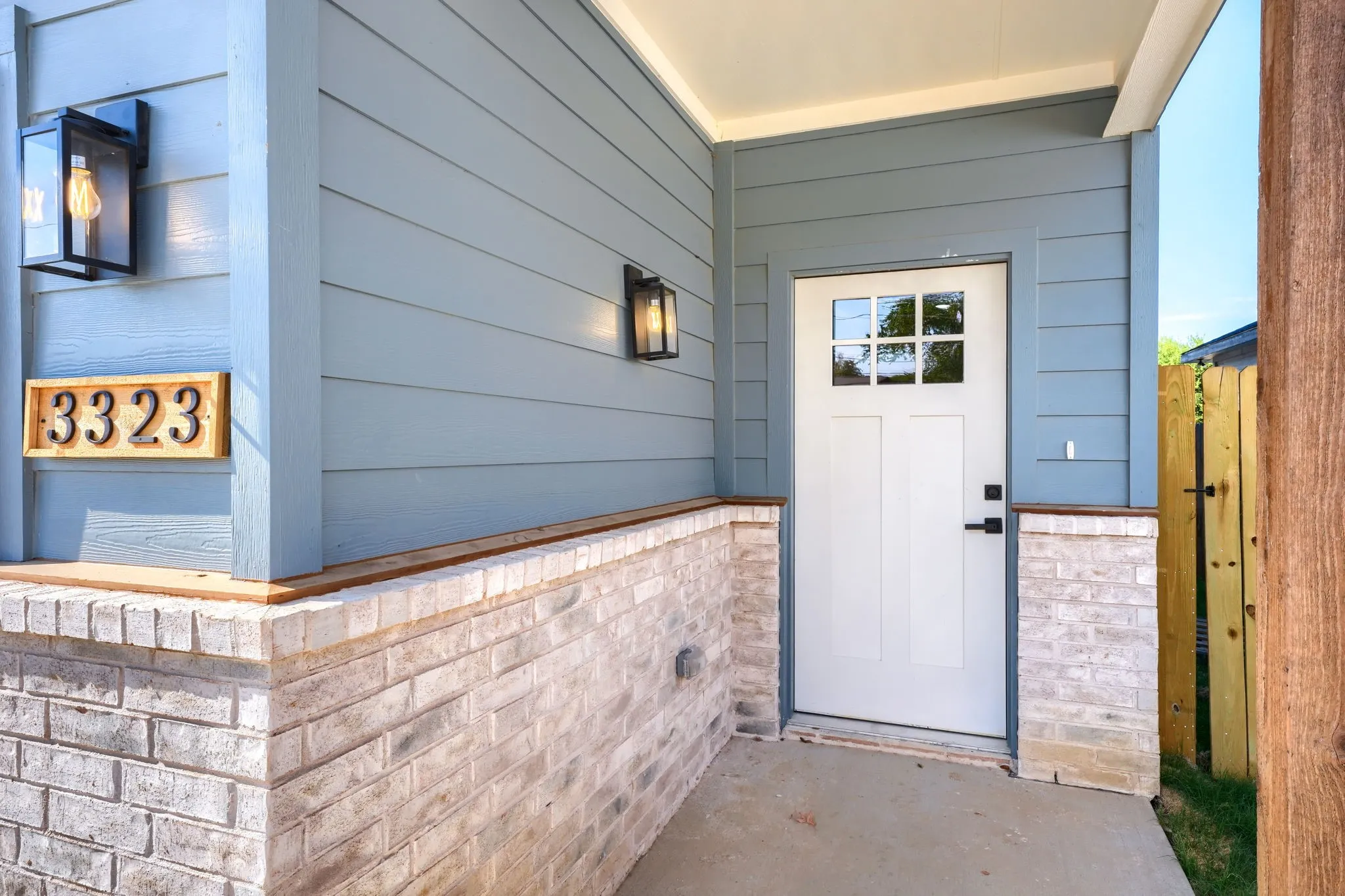 Front entry featuring light blue siding, brick detailing, modern exterior lighting, an address plaque, and a white paneled door with windowpanes