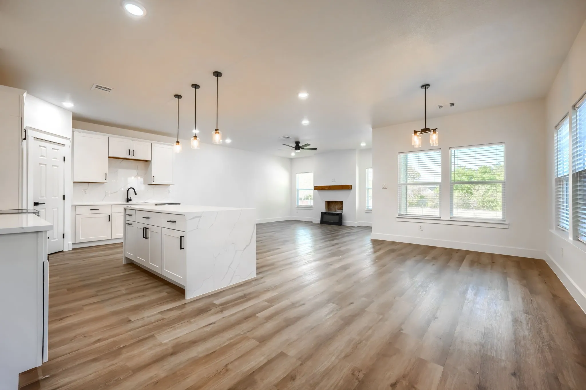 Open-concept layout featuring wood-finish flooring, a kitchen island with stone countertops, white cabinetry, and a fireplace with a wood mantle