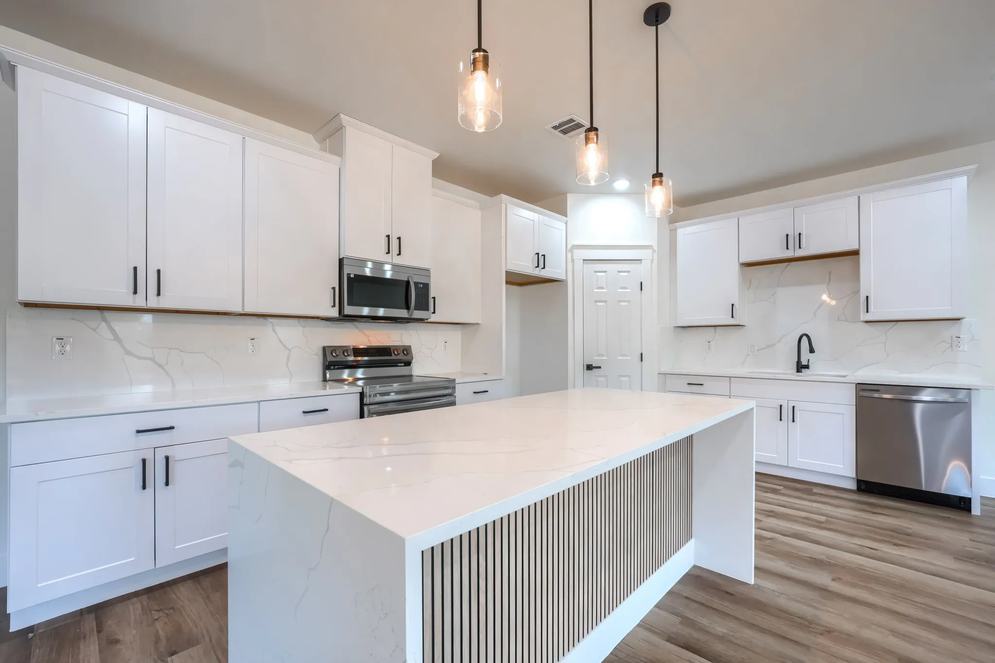White shaker cabinetry, light-toned wood-finish flooring, stainless steel appliances, white countertops with veining, and a kitchen island with slat-panel detailing