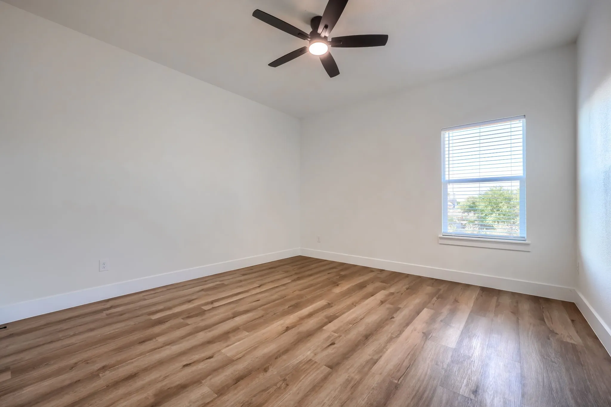 Wood-finish flooring, white walls, and a window with blinds, complemented by a ceiling fan