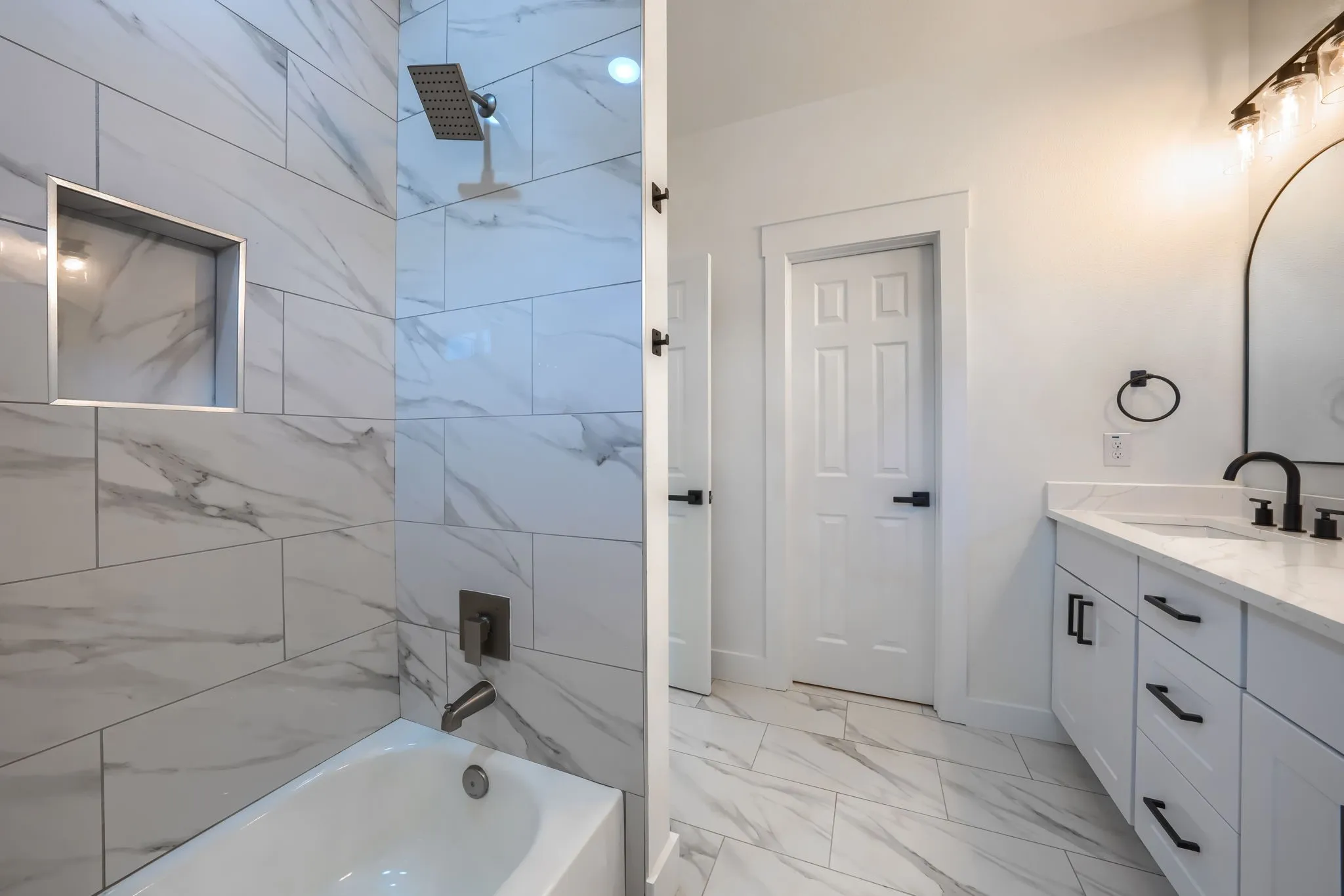 White tile flooring, white vanity with integrated sink, black matte fixtures, white subway tile shower surround with integrated niche, and a white bathtub
