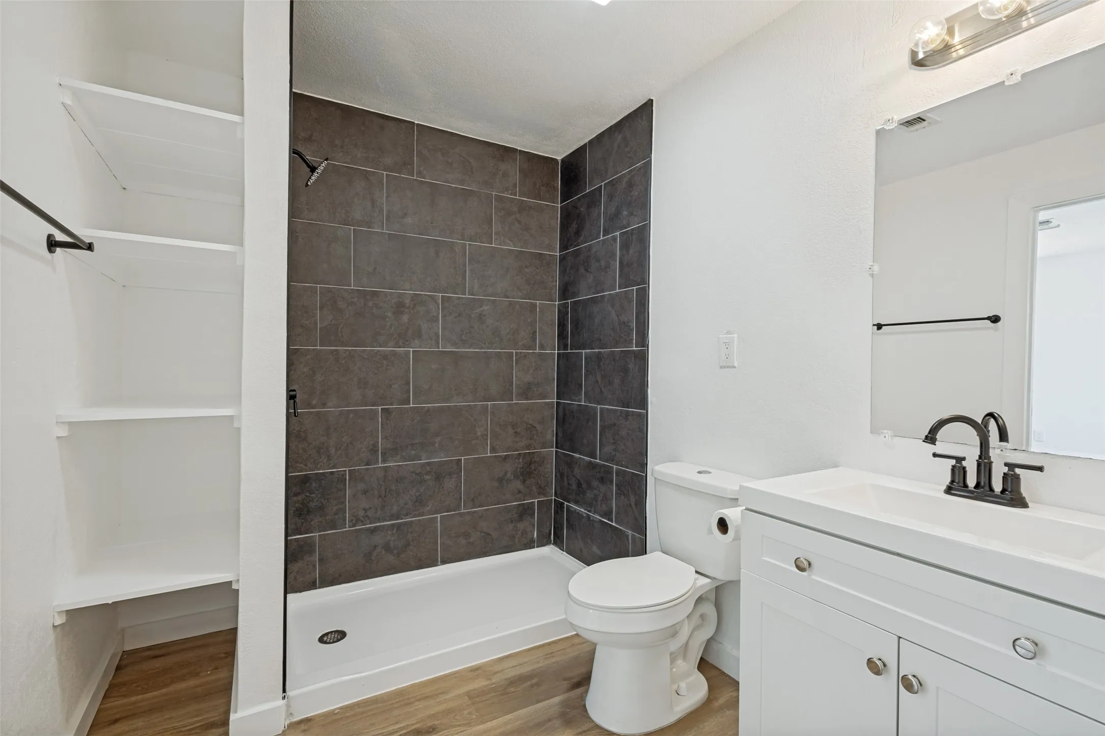 Bathroom featuring wood-finish flooring, a tiled shower surround, a white vanity with cabinet storage, and a wall-mounted mirror