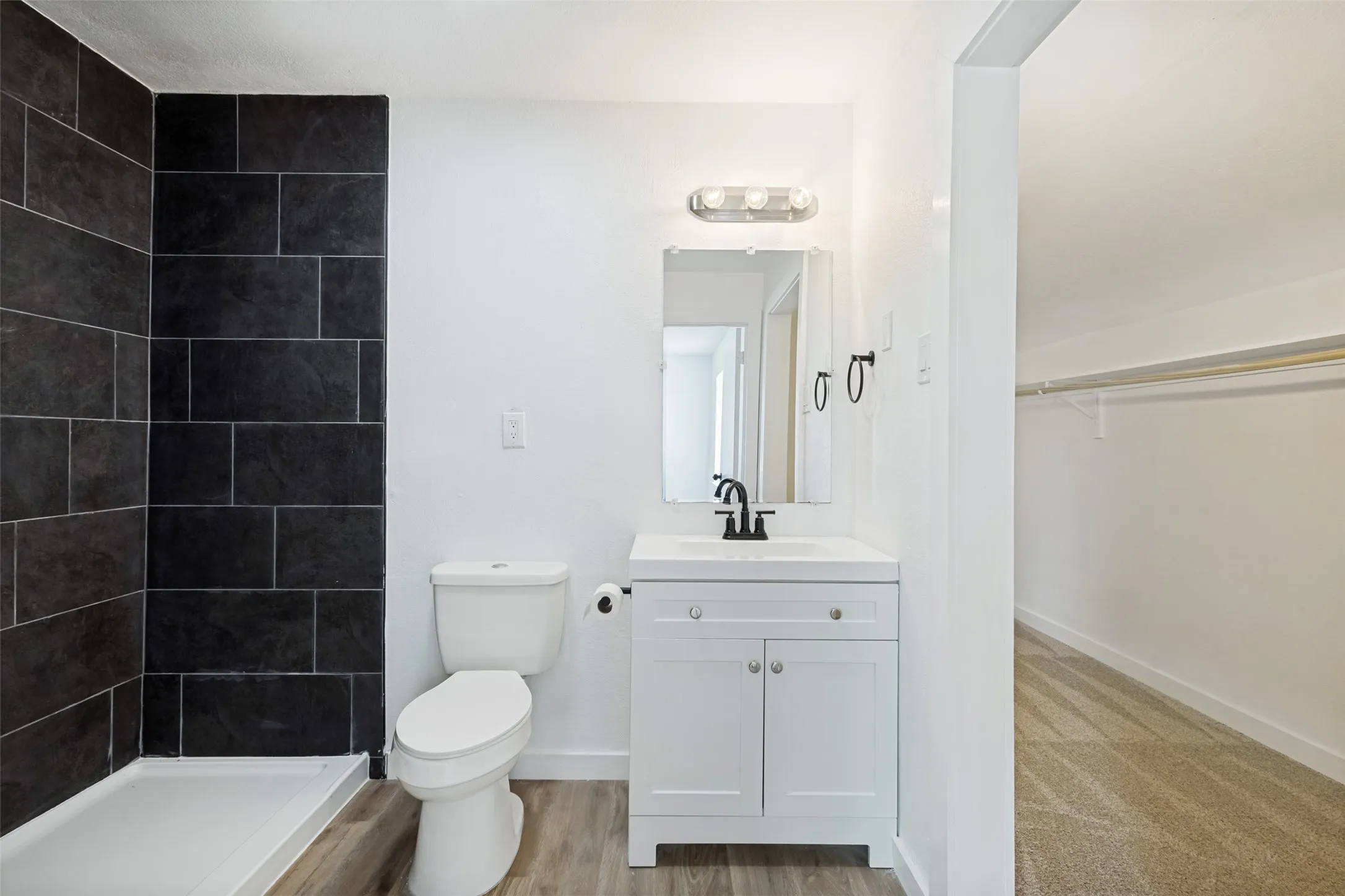 Bathroom featuring a walk-in shower with dark tile surround, a white vanity with integrated sink, and wood-finish flooring