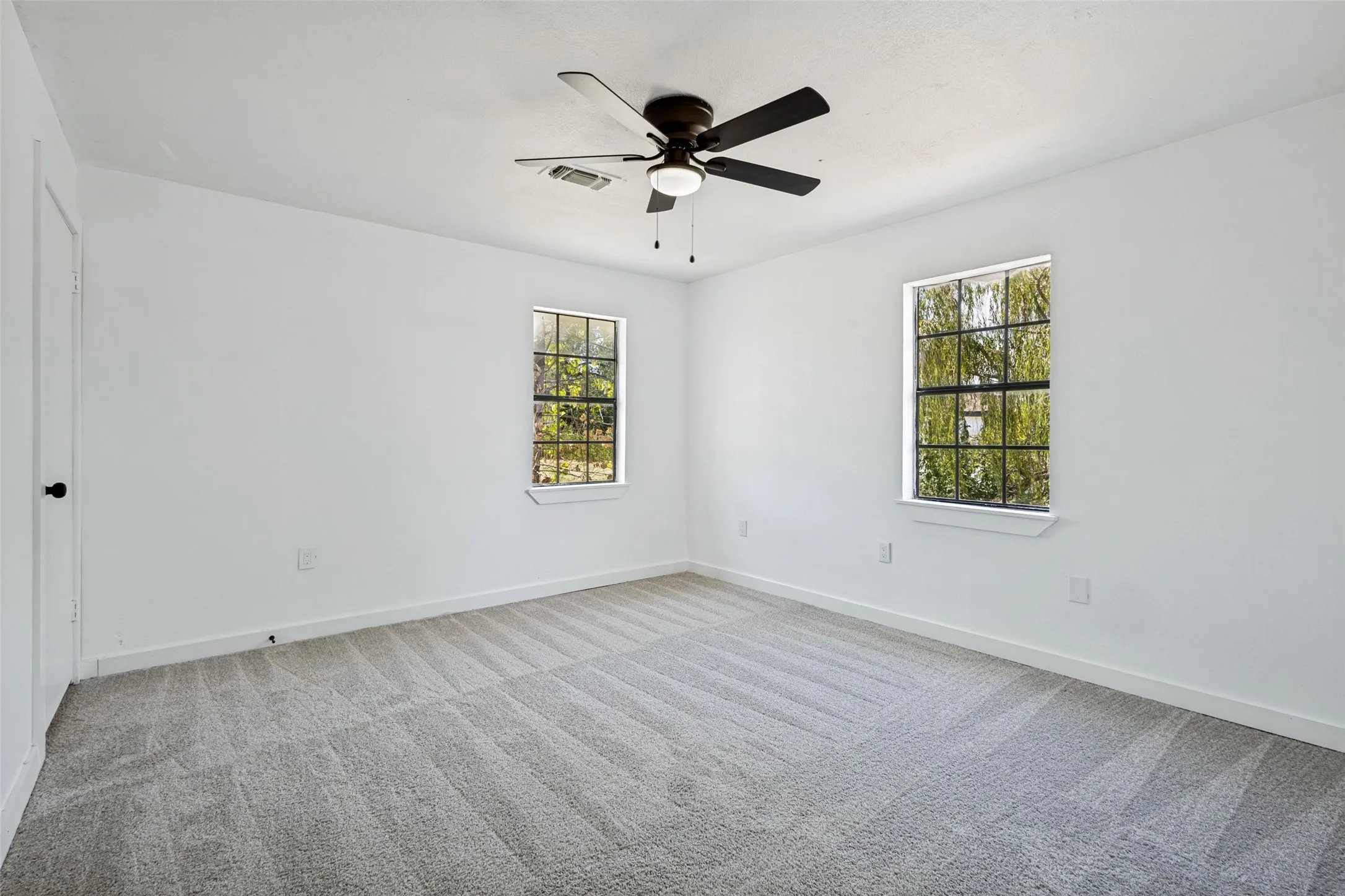 Light gray patterned carpeting, a dark bronze ceiling fan with integrated lighting, and two windows with white trim