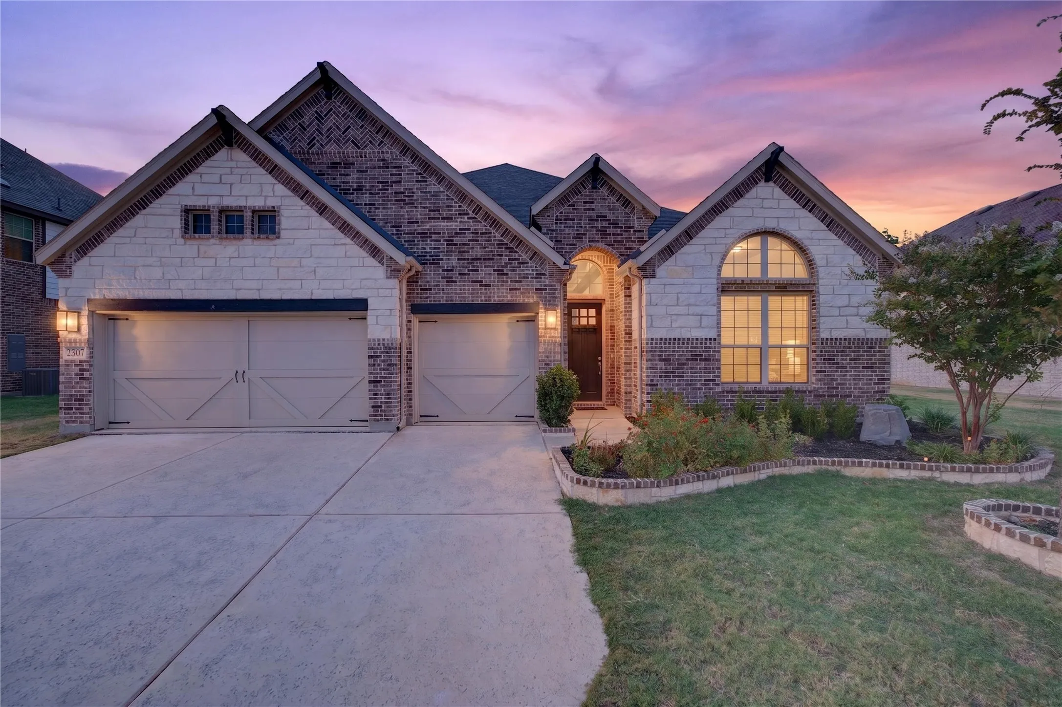 Brick and stone exterior with arched windows, a two-car garage, and landscaped garden beds