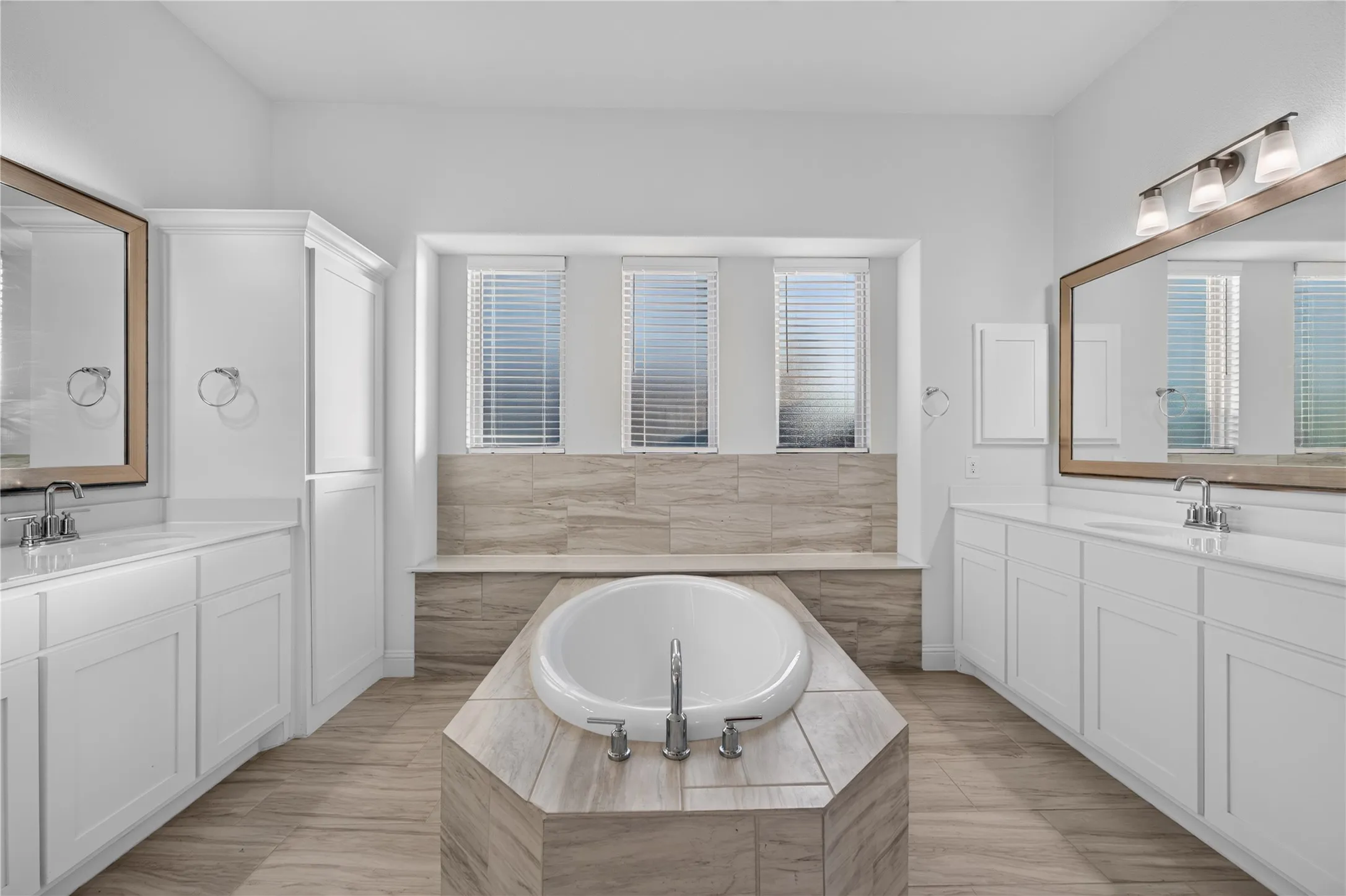 Freestanding soaking tub with tiled surround, dual vanities with white cabinetry, framed mirrors, and wood-finish flooring