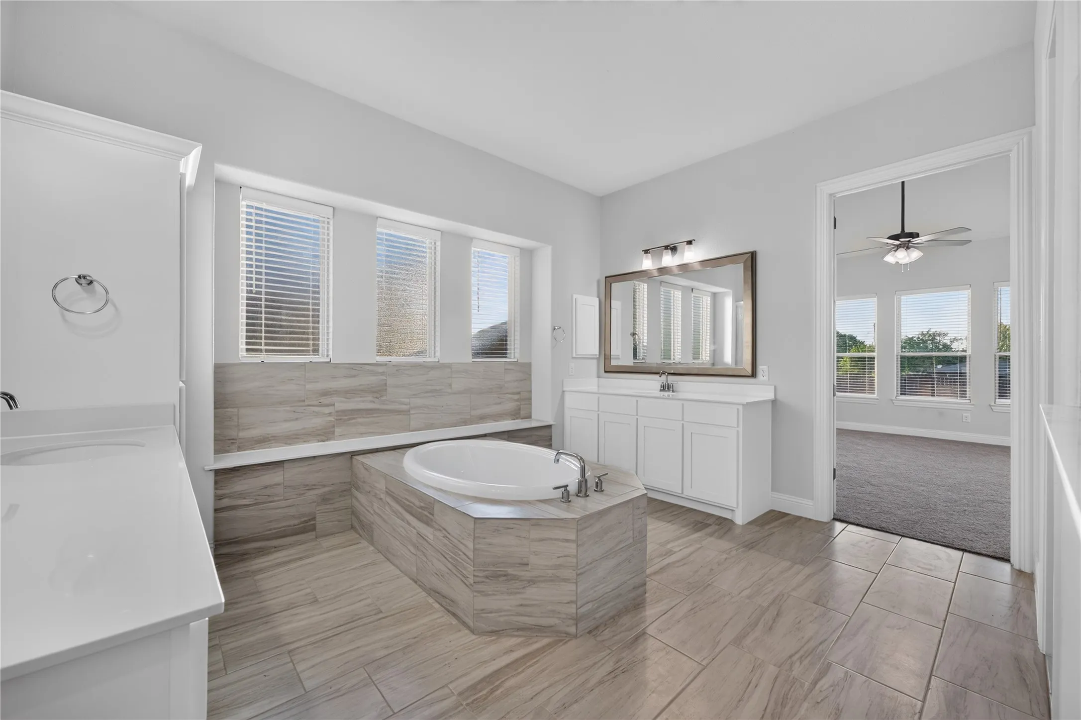 Built-in soaking tub with surrounding tiled platform, dual vanities with white cabinetry and solid surface countertops, tiled flooring, and multiple windows with blinds