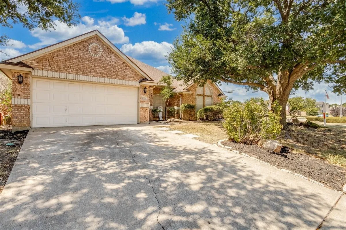 Brick exterior with an attached two-car garage, concrete driveway, mature trees, and landscaped shrubbery