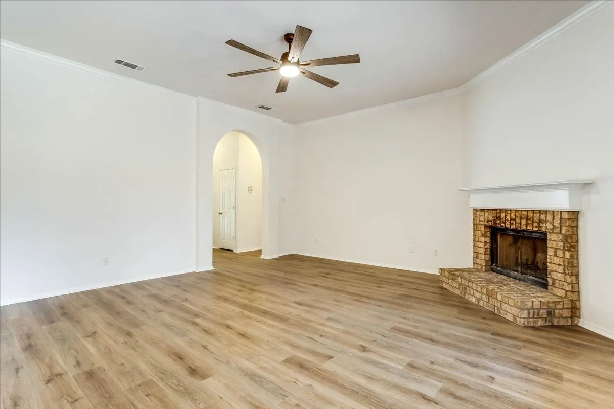 Wood-finish flooring, a brick fireplace with a white mantel, an arched entryway, and a ceiling fan with light fixture