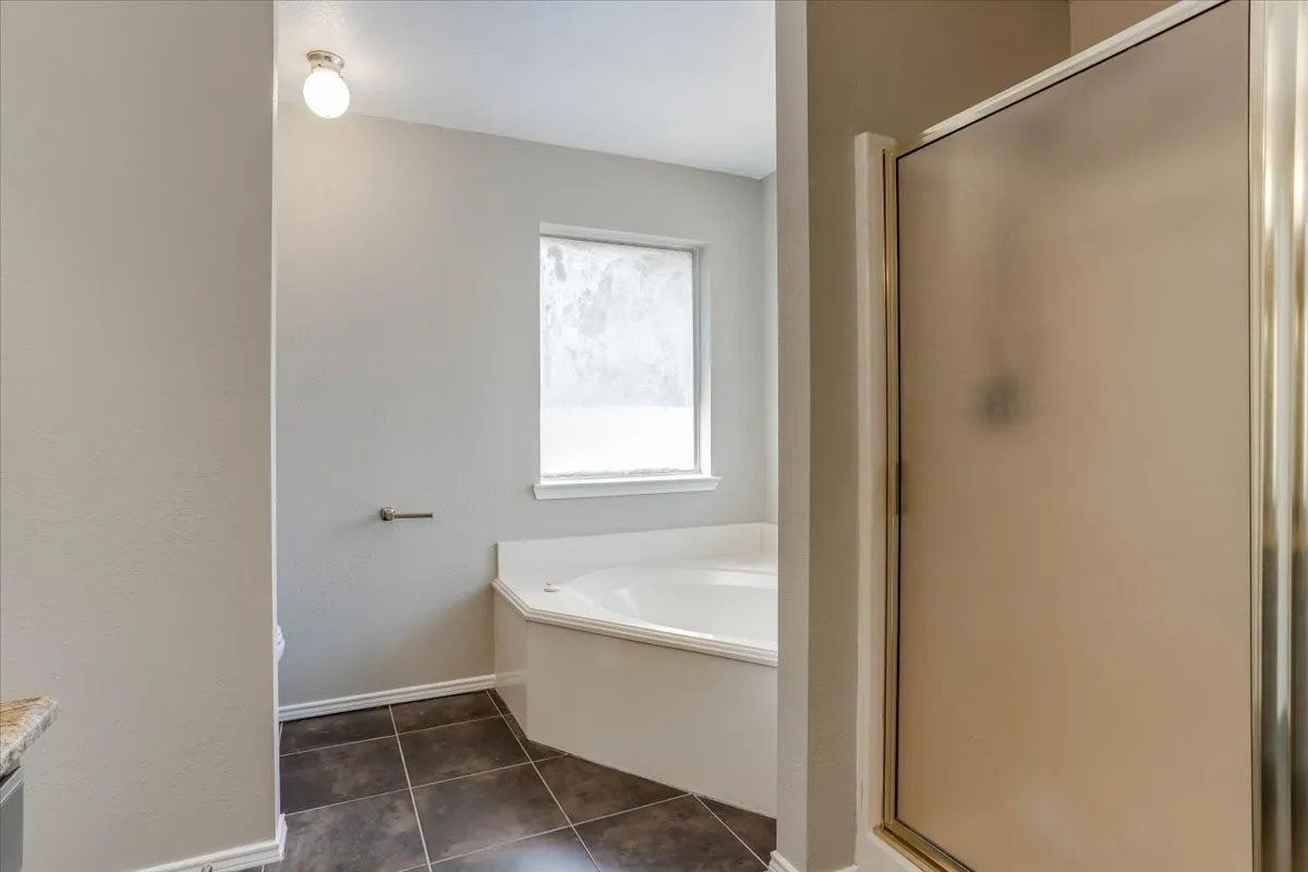 Corner soaking tub with a window, tile flooring, and an enclosed shower with a frosted door