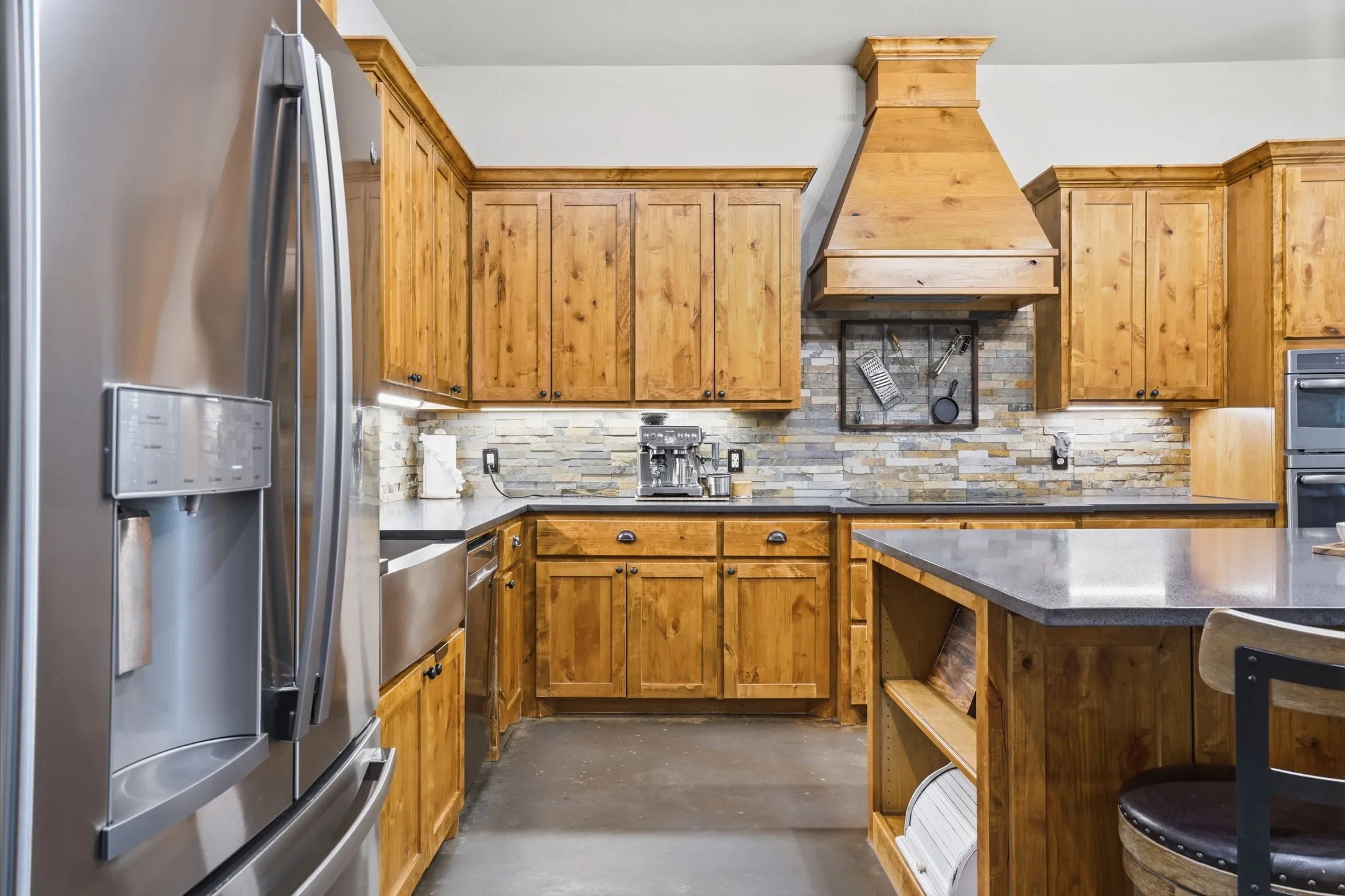 Stainless steel refrigerator, custom wood cabinetry, stone-tiled backsplash, built-in range hood, and dark countertops
