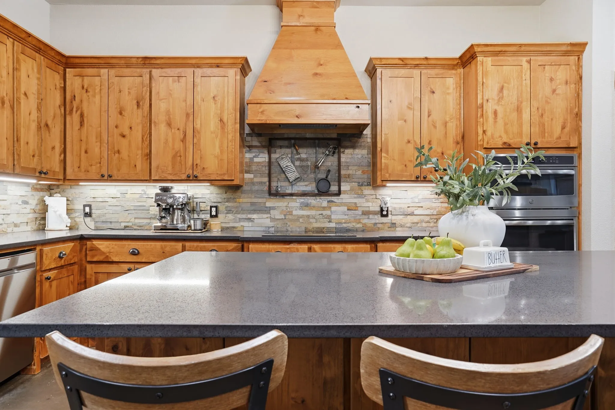 Spacious kitchen featuring a large island with dark countertops, wood cabinetry, built-in double ovens, and a stone-finish backsplash
