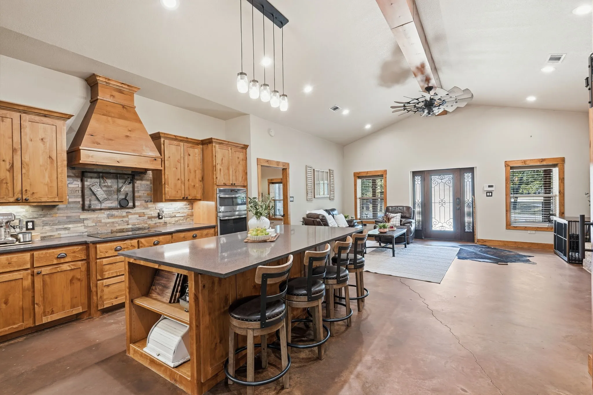 Open-concept living area featuring a kitchen island with seating, wood-finish cabinetry, stainless steel double ovens, a vaulted ceiling, and a decorative front door with sidelights