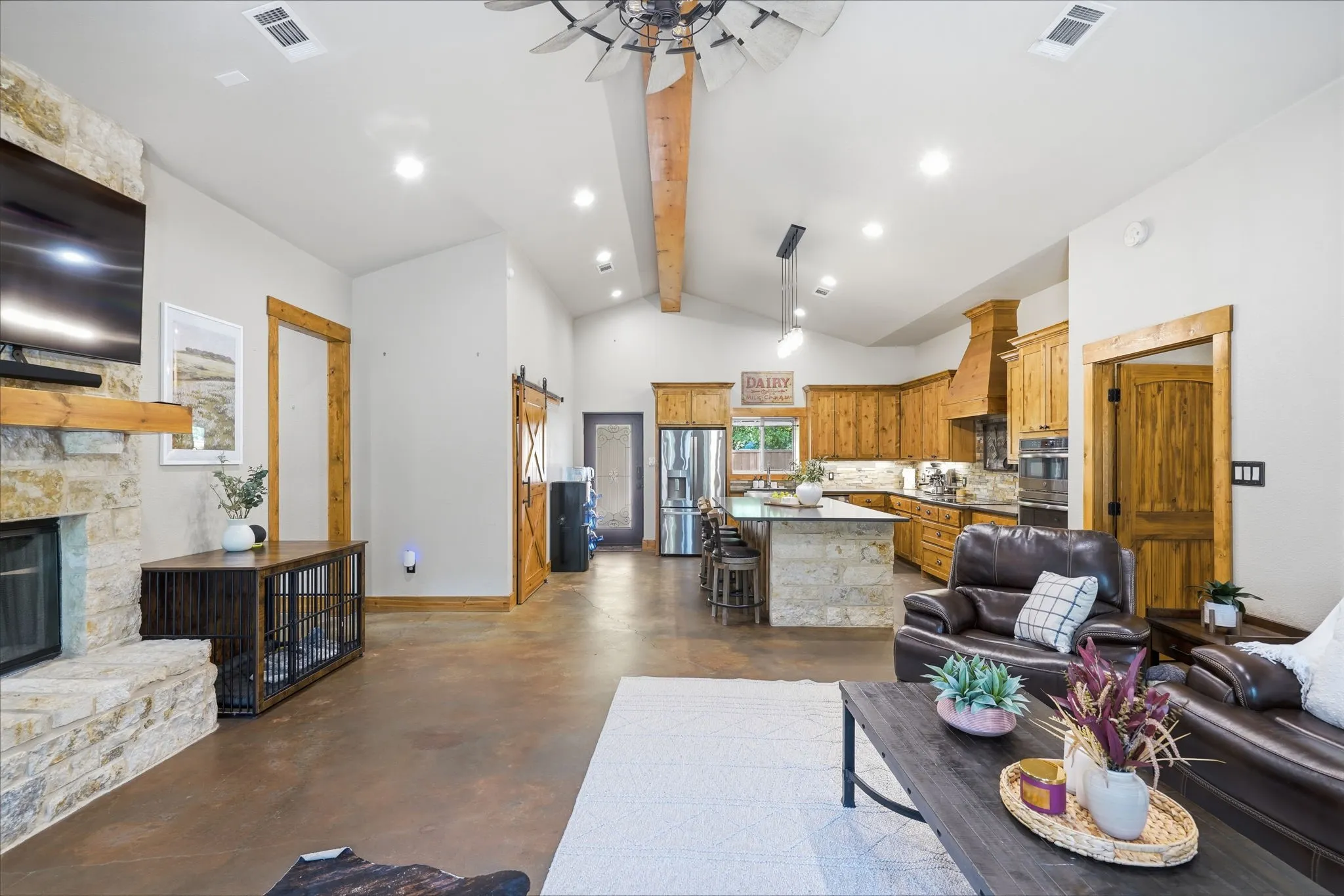 Vaulted ceilings featuring exposed wood beams, a stone fireplace, wood-finish flooring, a kitchen island with stone-clad base, and built-in wood cabinetry