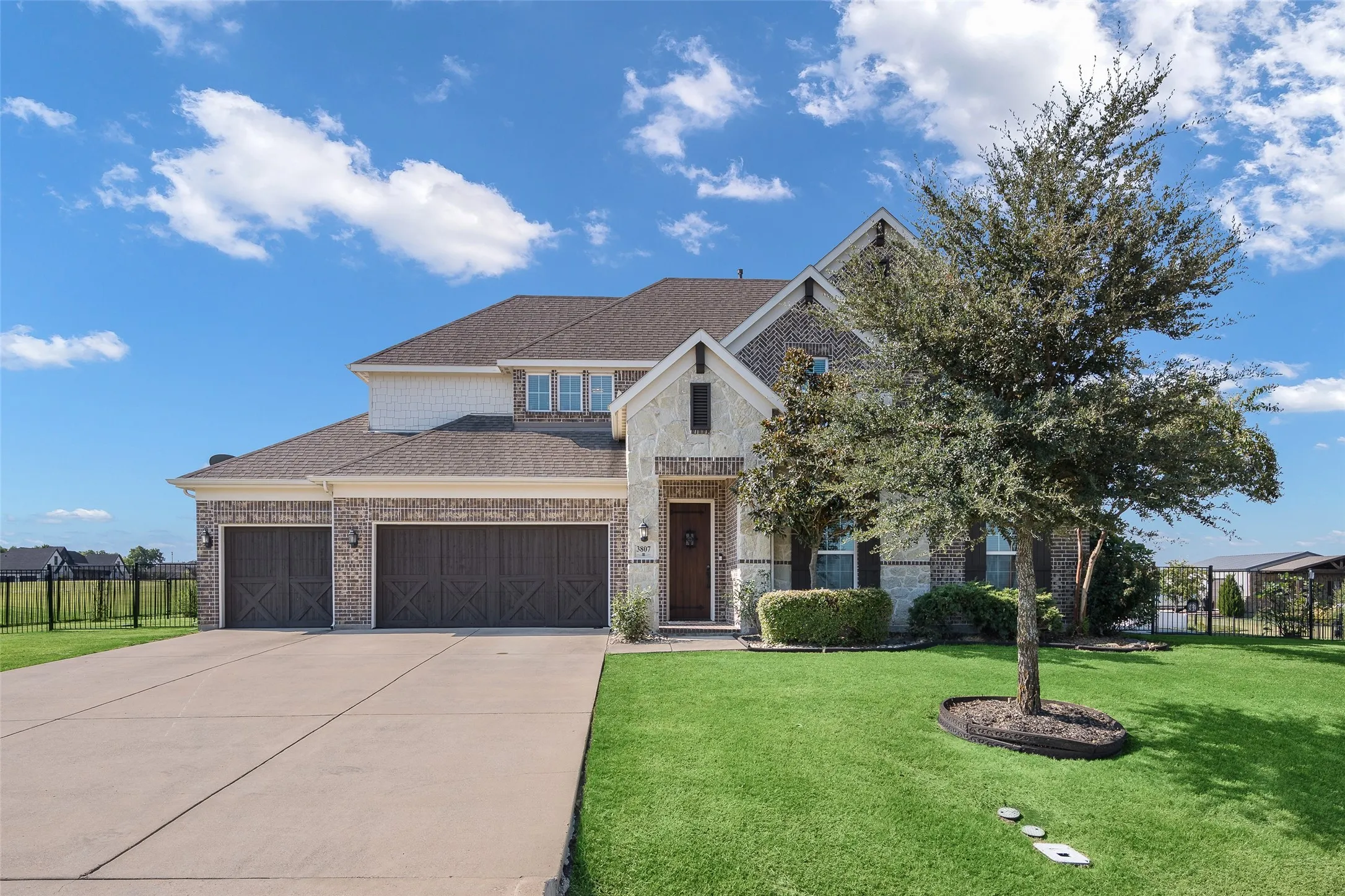 Stone and brick facade with multiple gables, three-car garage, concrete driveway, manicured lawn, and mature tree