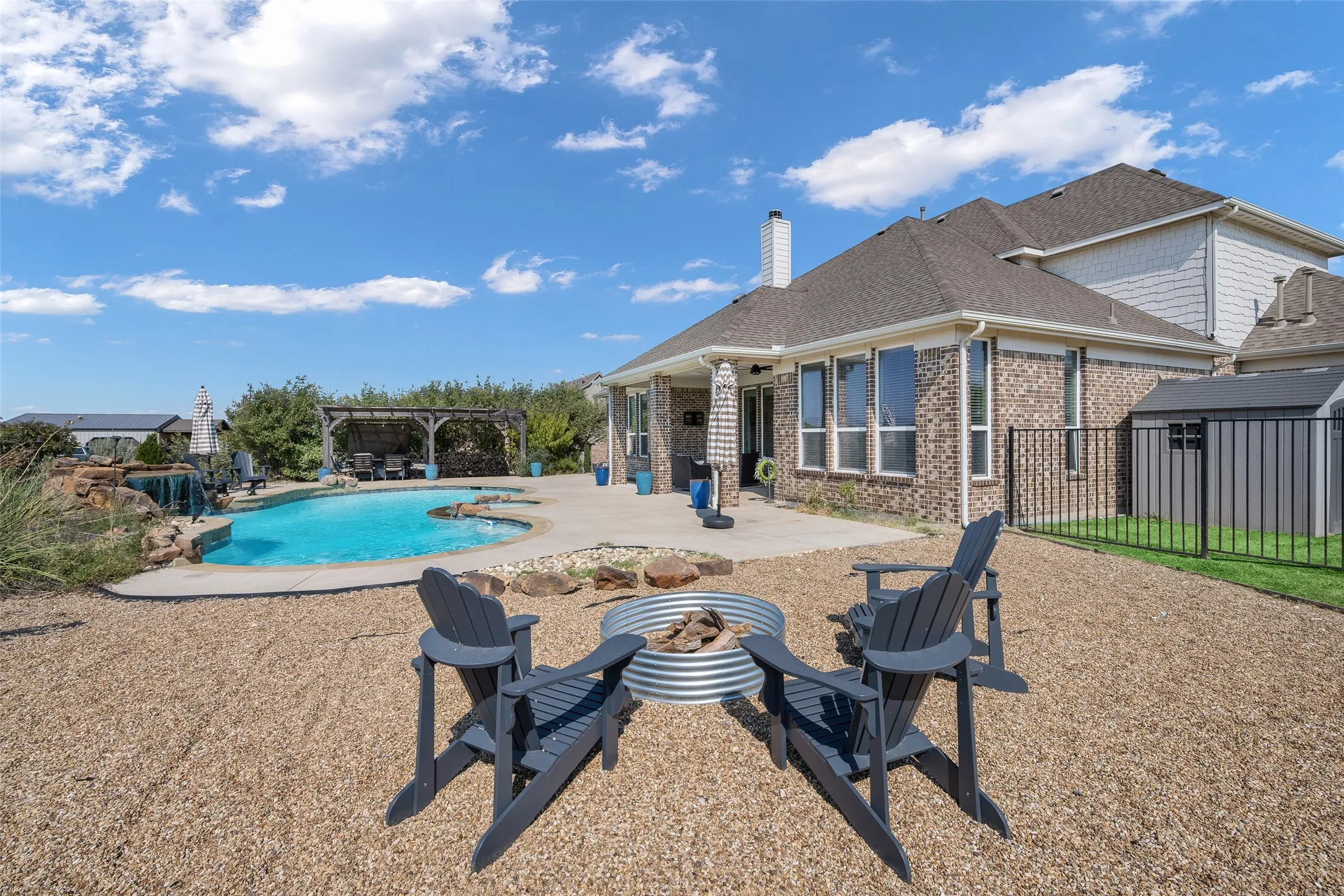 Expansive backyard featuring a built-in pool with a rock waterfall, an integrated spa, a dedicated gravel fire pit area, and an outdoor pergola