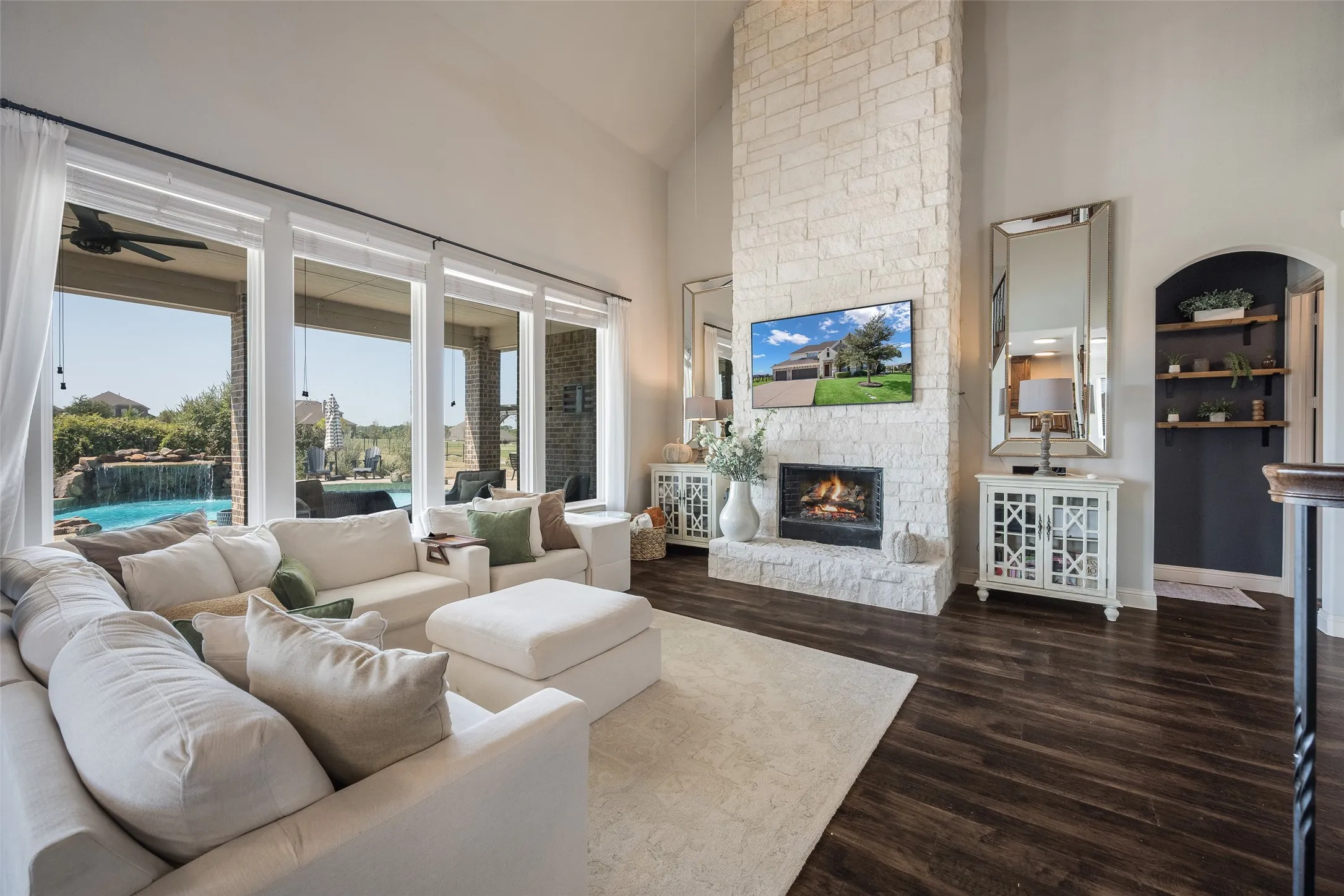 Vaulted ceiling with tall windows, stone-clad fireplace, dark wood-finish flooring, and an arched entryway with built-in shelving