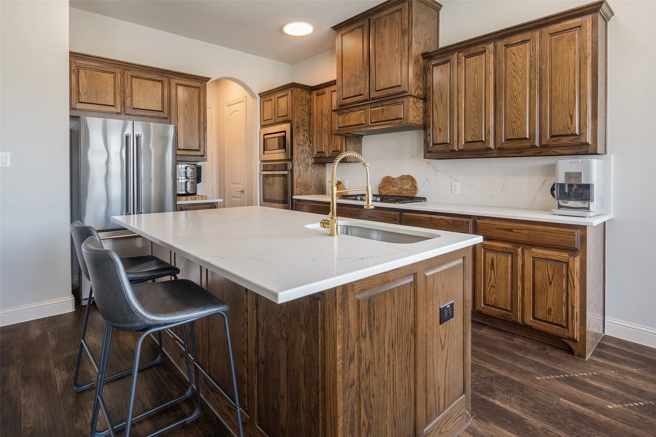 Kitchen featuring wood-finish cabinetry, stainless steel appliances, a large island with a surface-mount sink, and light-toned countertops with a matching backsplash