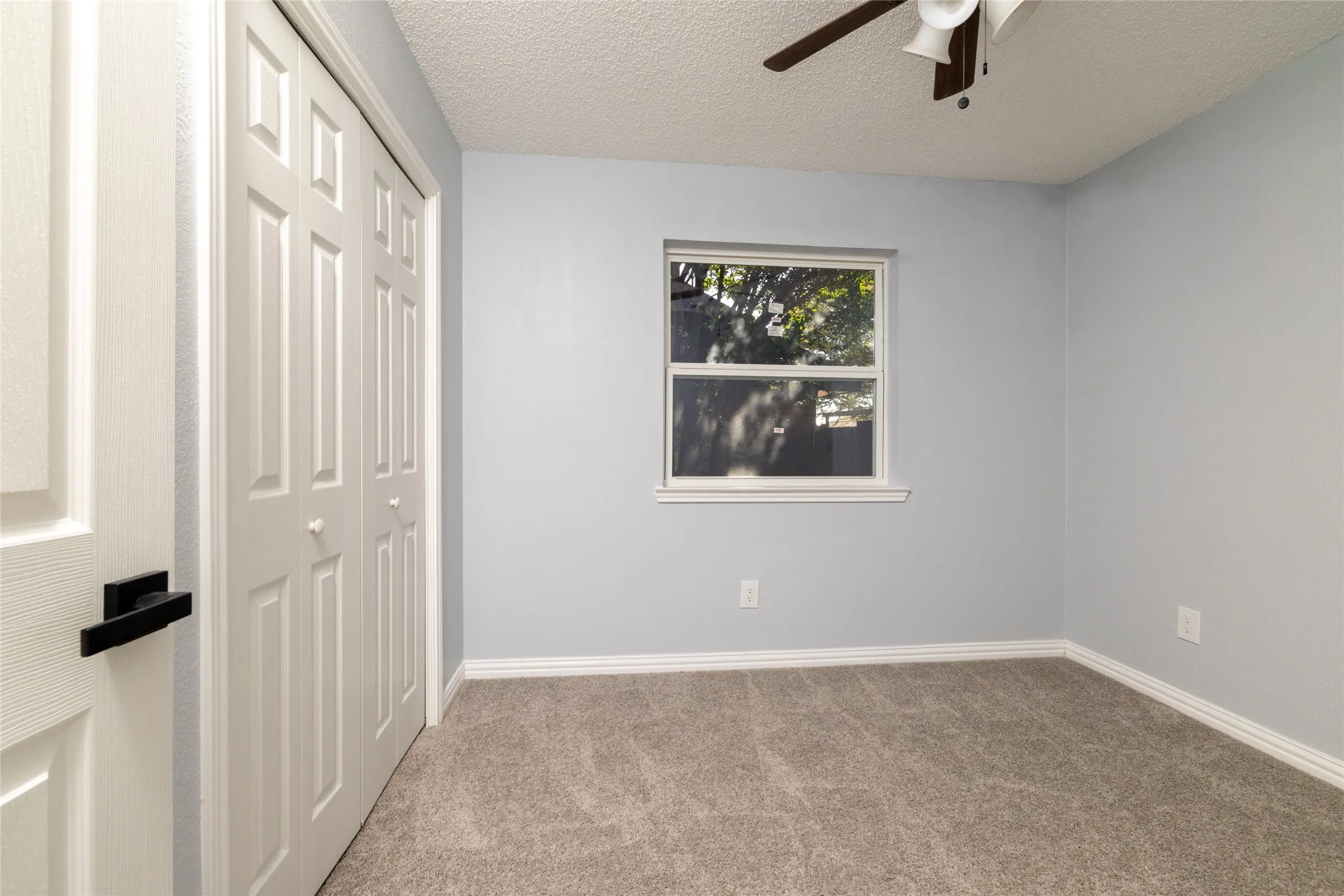 Light gray wall paint, textured ceiling, ceiling fan with light fixture, window, and gray carpet flooring