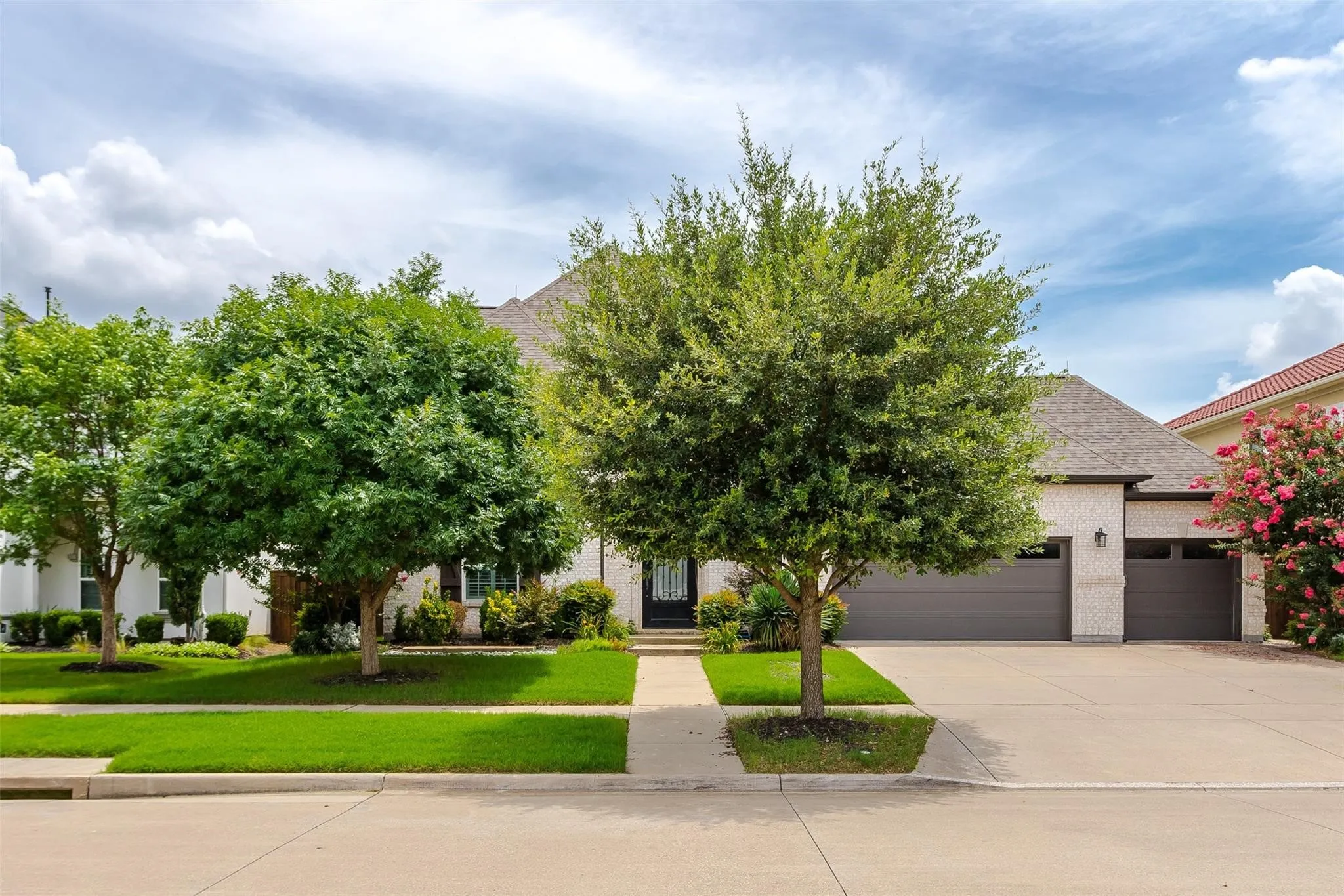 Exterior featuring a light brick facade, dual garage doors, a concrete driveway, a well-maintained lawn, and mature trees
