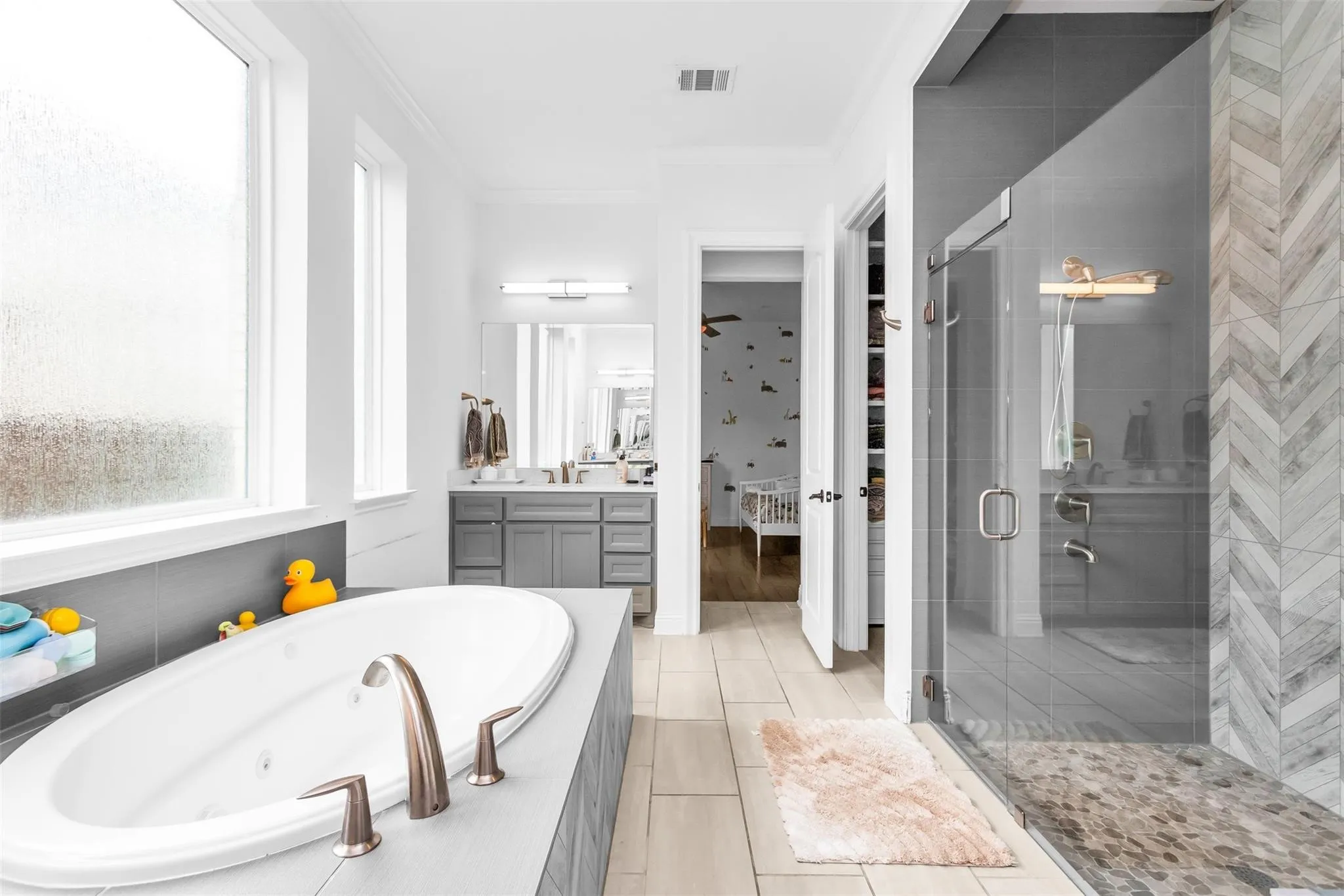 Built-in soaking tub with bronze-finish fixtures, a walk-in shower with glass enclosure, chevron-patterned tile, and a dual vanity with gray cabinetry