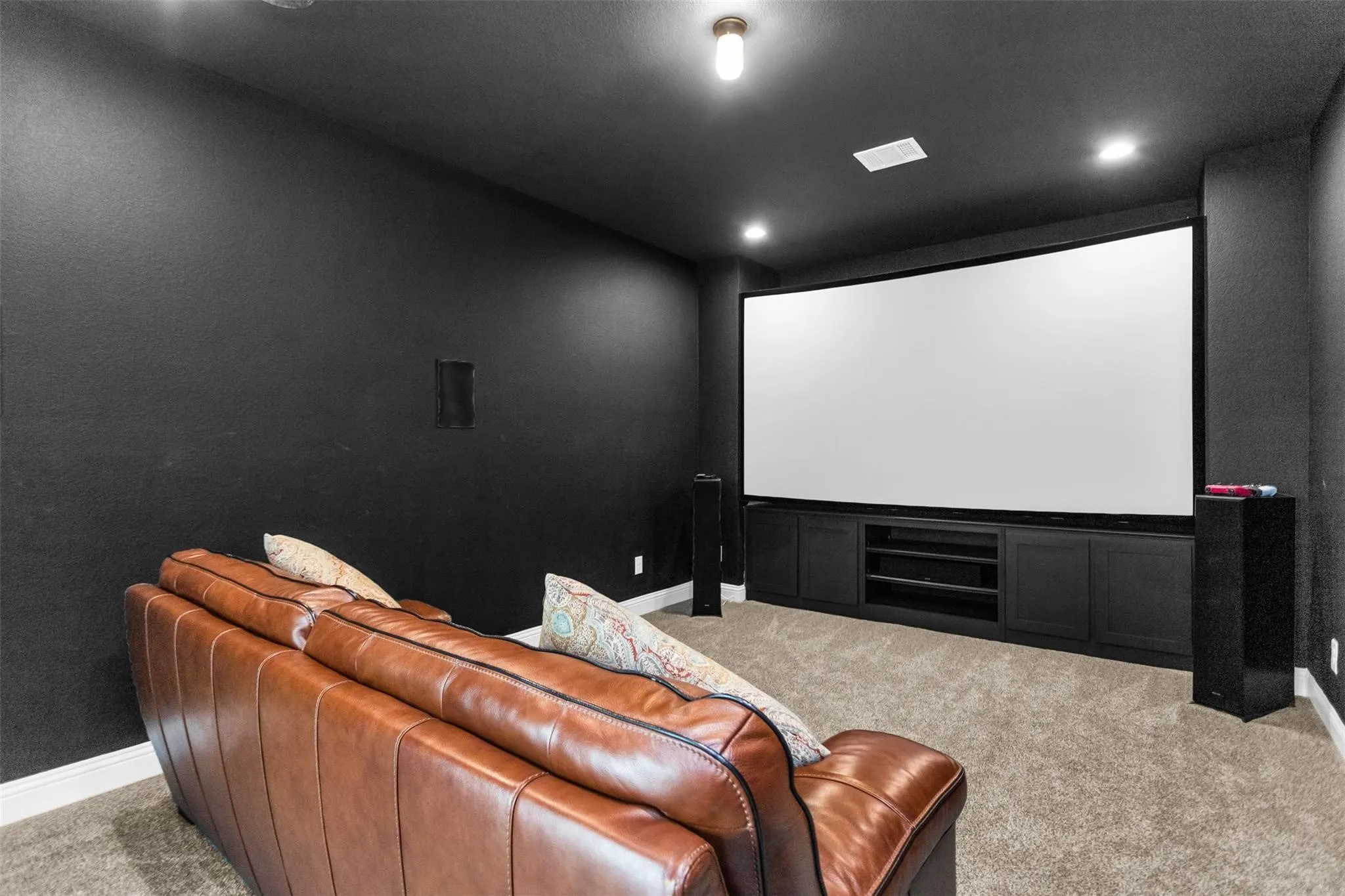 Dedicated media room featuring a large curved projection screen, built-in media console with storage, dark walls, and recessed lighting