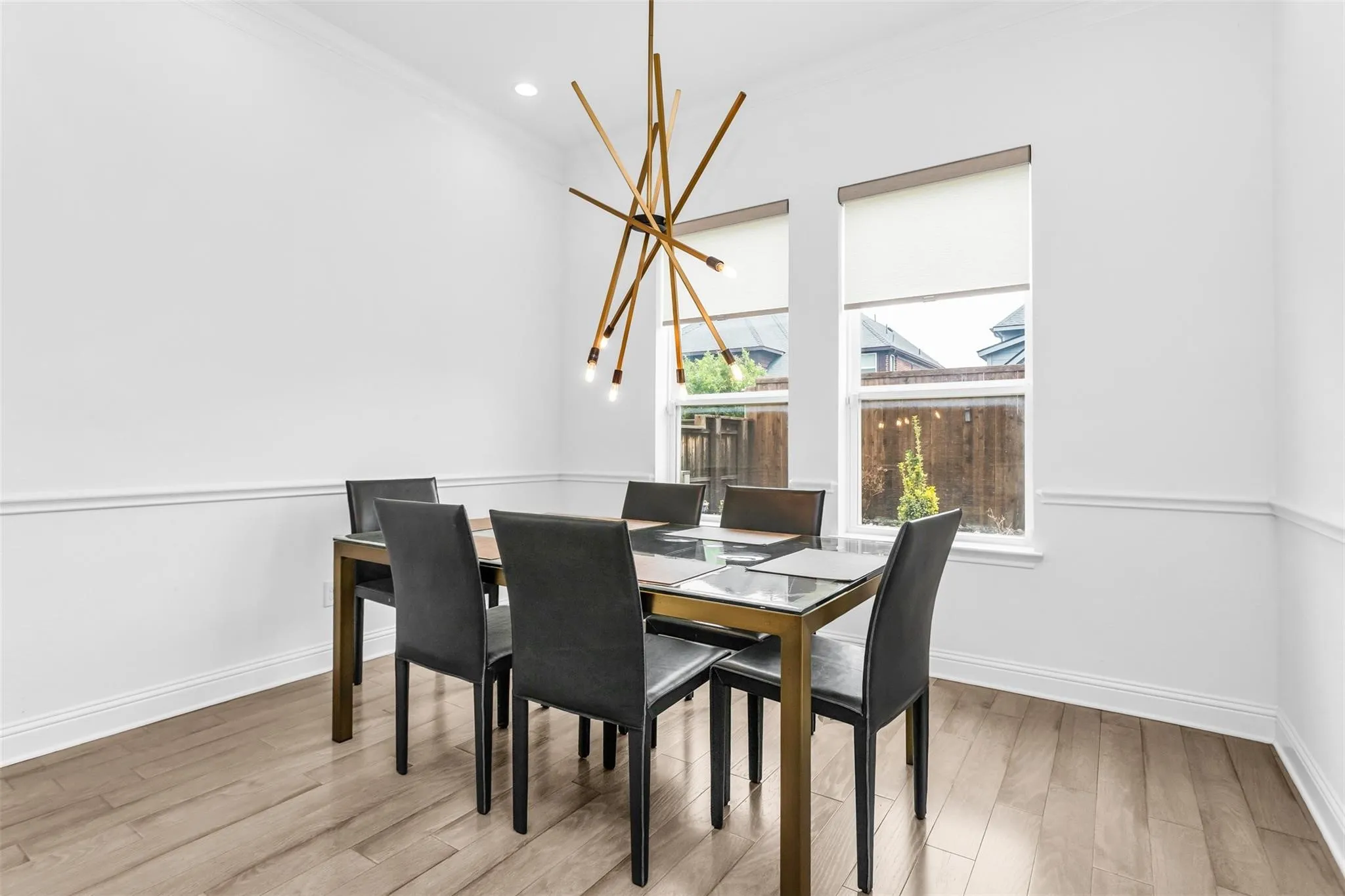 Wood-finish flooring, white walls with decorative molding, modern chandelier, recessed lighting, and two windows with roller shades