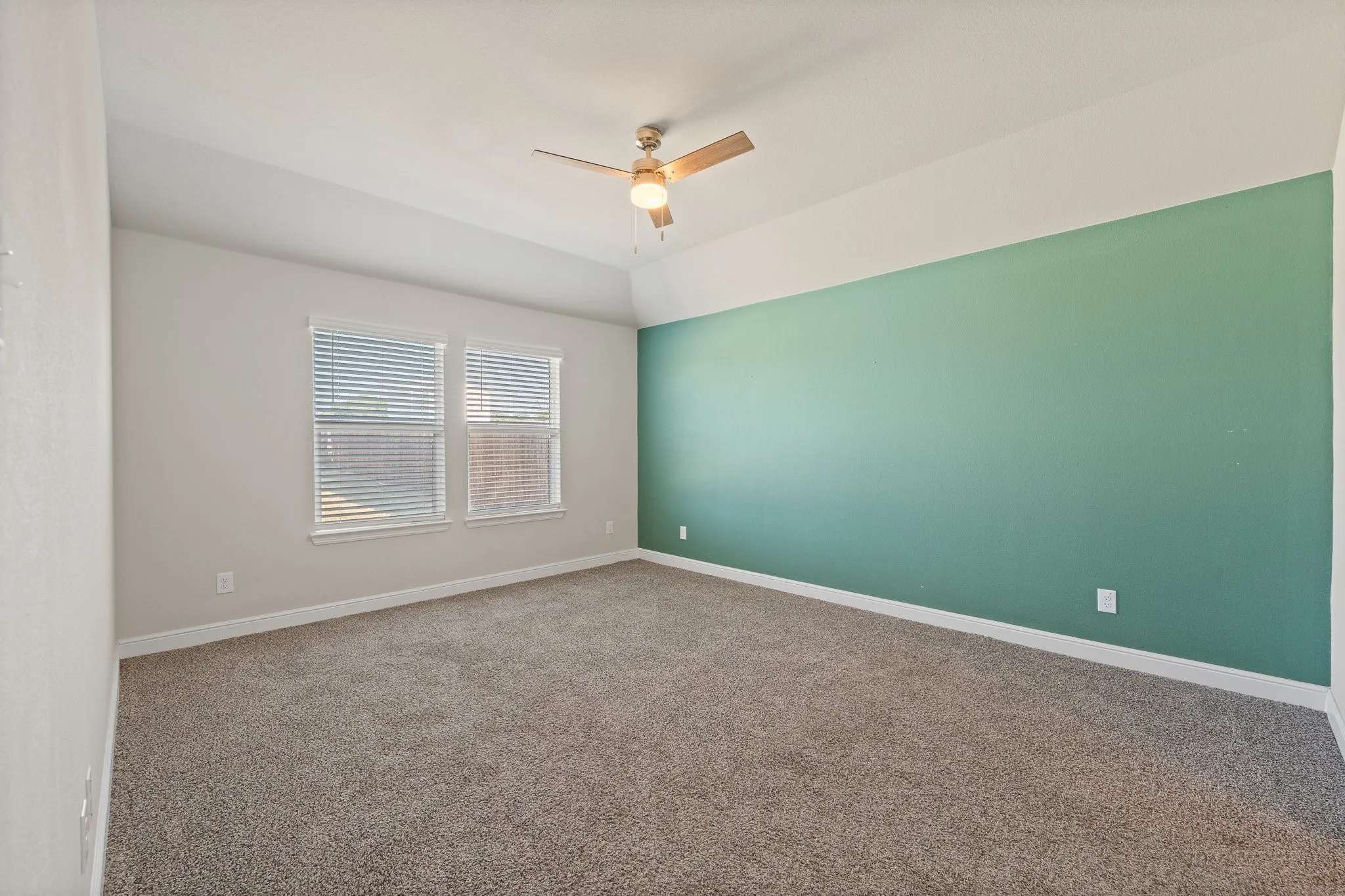 Vaulted ceiling, two windows with blinds, ceiling fan with integrated lighting, carpet flooring, accent wall