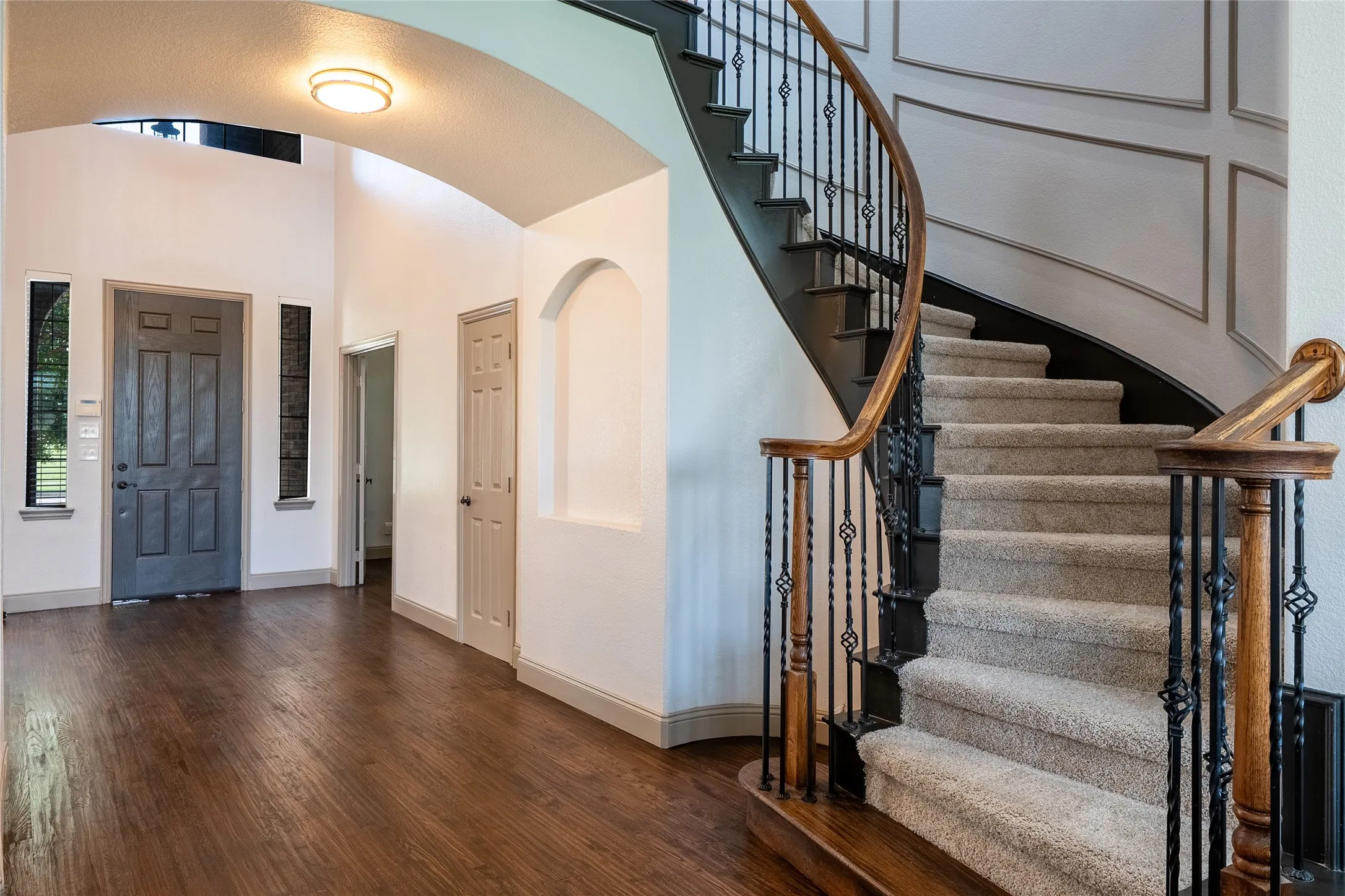 Wood-finish flooring, grand curved staircase with wrought iron balusters, arched entryway, architectural wall paneling, and an arched wall niche