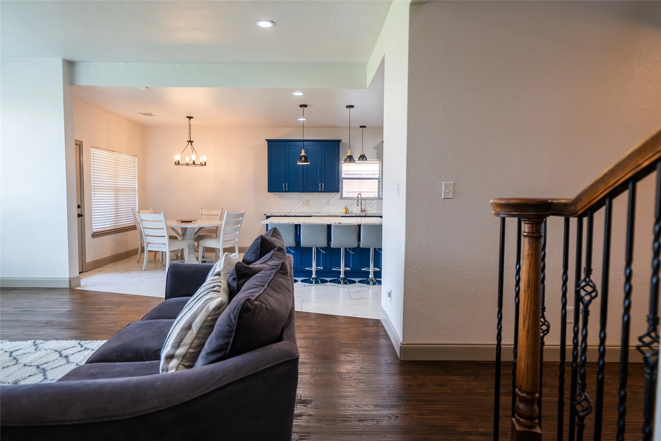 Wood-finish flooring, staircase with wrought iron balusters, recessed lighting, kitchen with blue cabinetry, and a dining area with a chandelier
