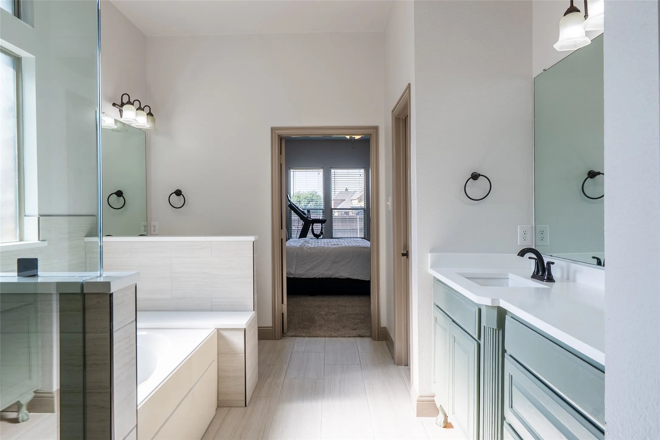 Dual vanity with white countertops, light wood-finish tile flooring, and a built-in bathtub with tiled surround