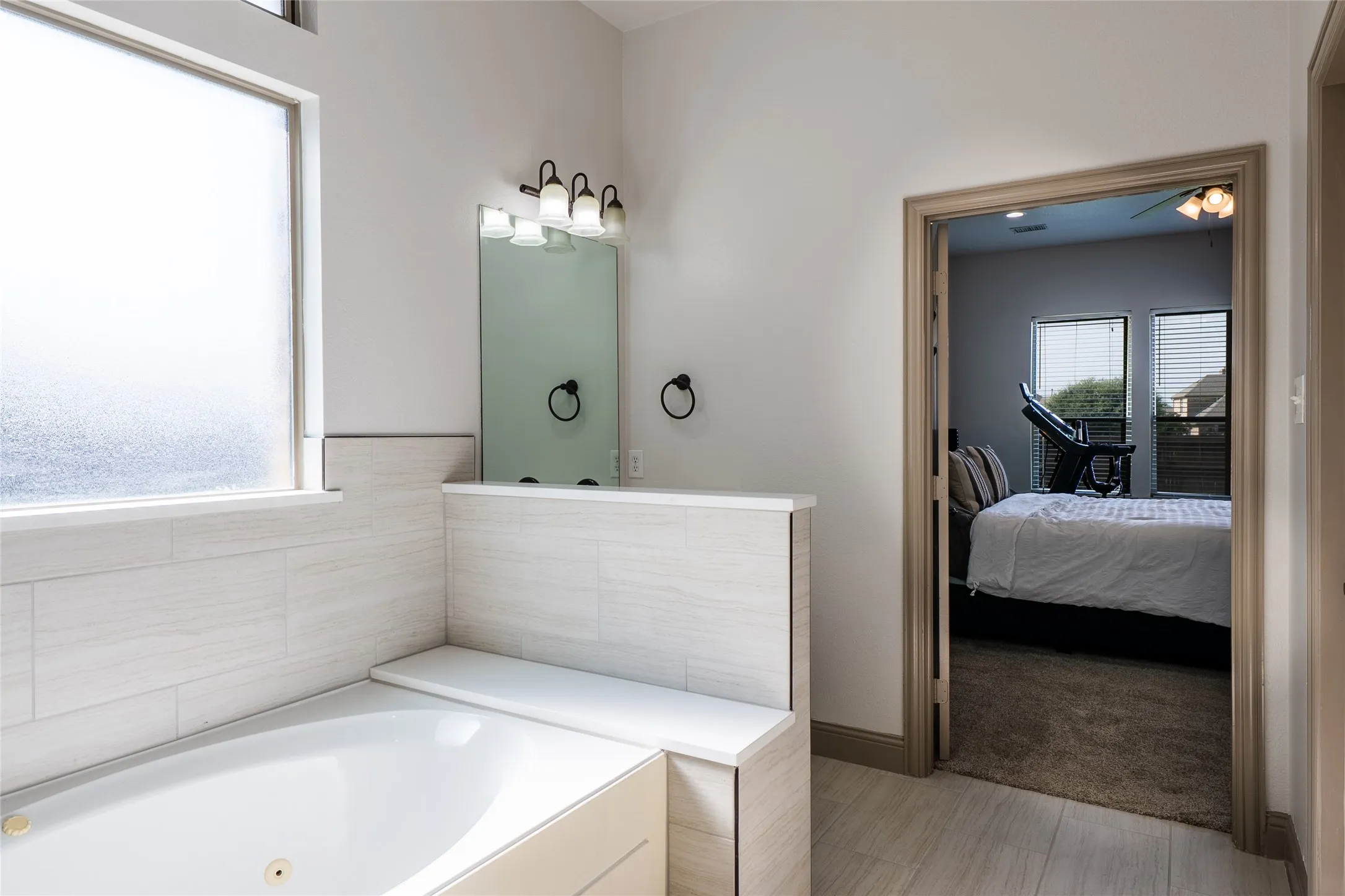 Jetted soaking tub with a tiled surround, frosted window, and an adjacent vanity with an integrated mirror and sconce lighting