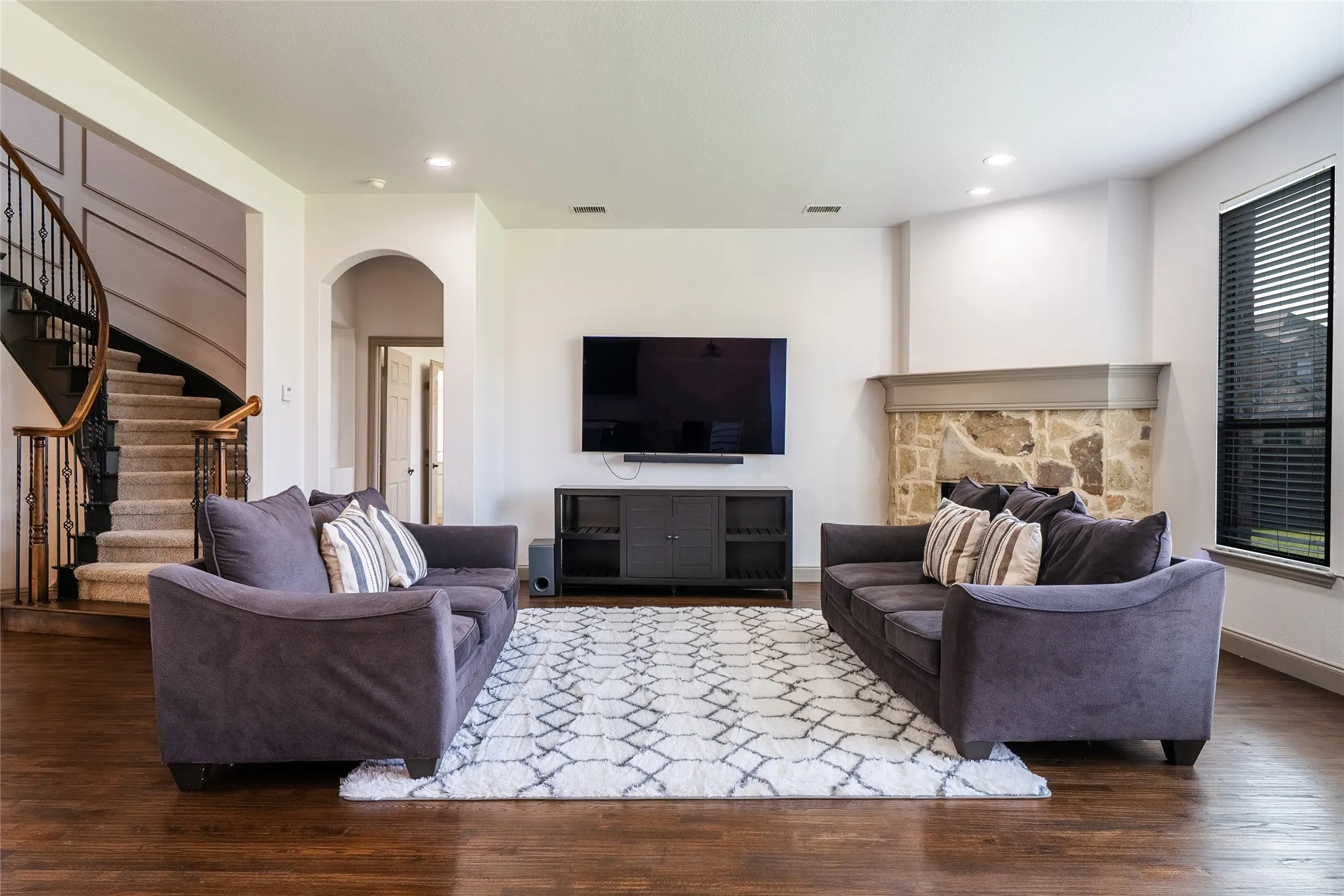 Wood-finish flooring, recessed lighting, stone-faced fireplace, large window with blinds, and a curving staircase with iron balusters