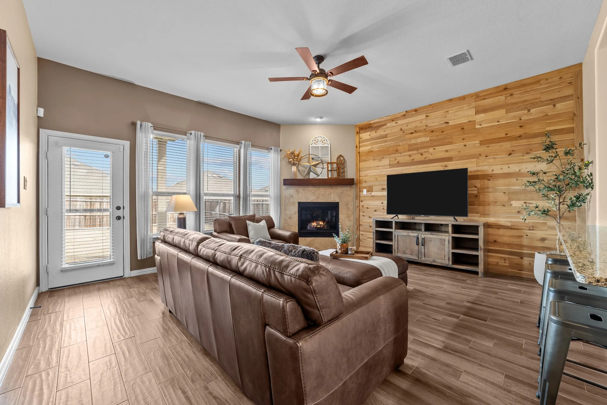 Living area featuring wood-finish flooring, a wood plank accent wall, a corner fireplace with a stone surround, and multiple windows with blinds