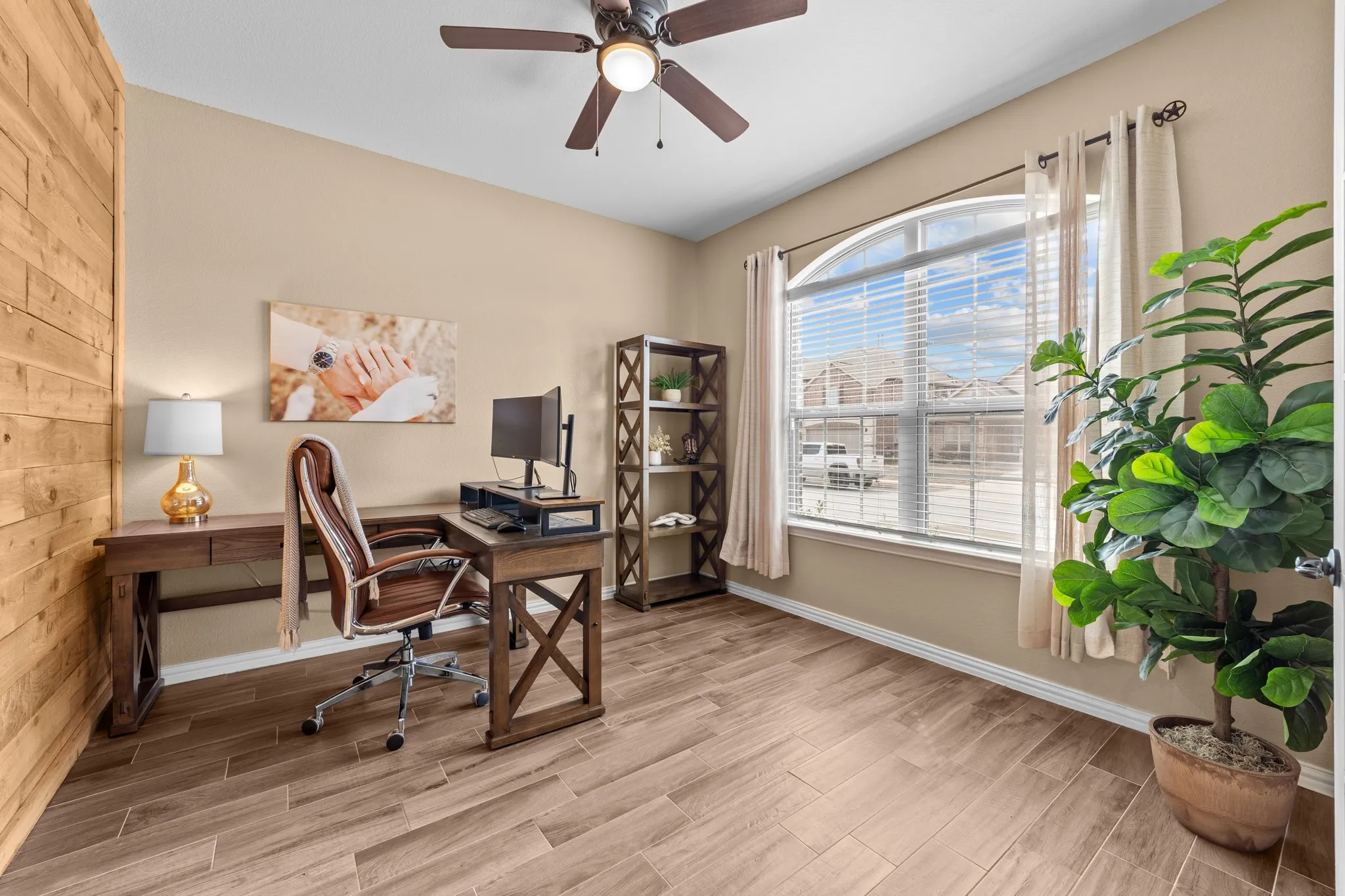 Wood-finish flooring, wood plank accent wall, arched window with horizontal blinds, ceiling fan with integrated lighting, and neutral wall tones