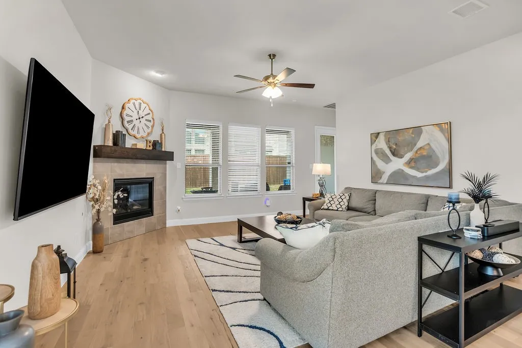 Wood-finish flooring, corner fireplace with tile surround, wood mantel, ceiling fan with integrated lighting, and multiple windows with blinds