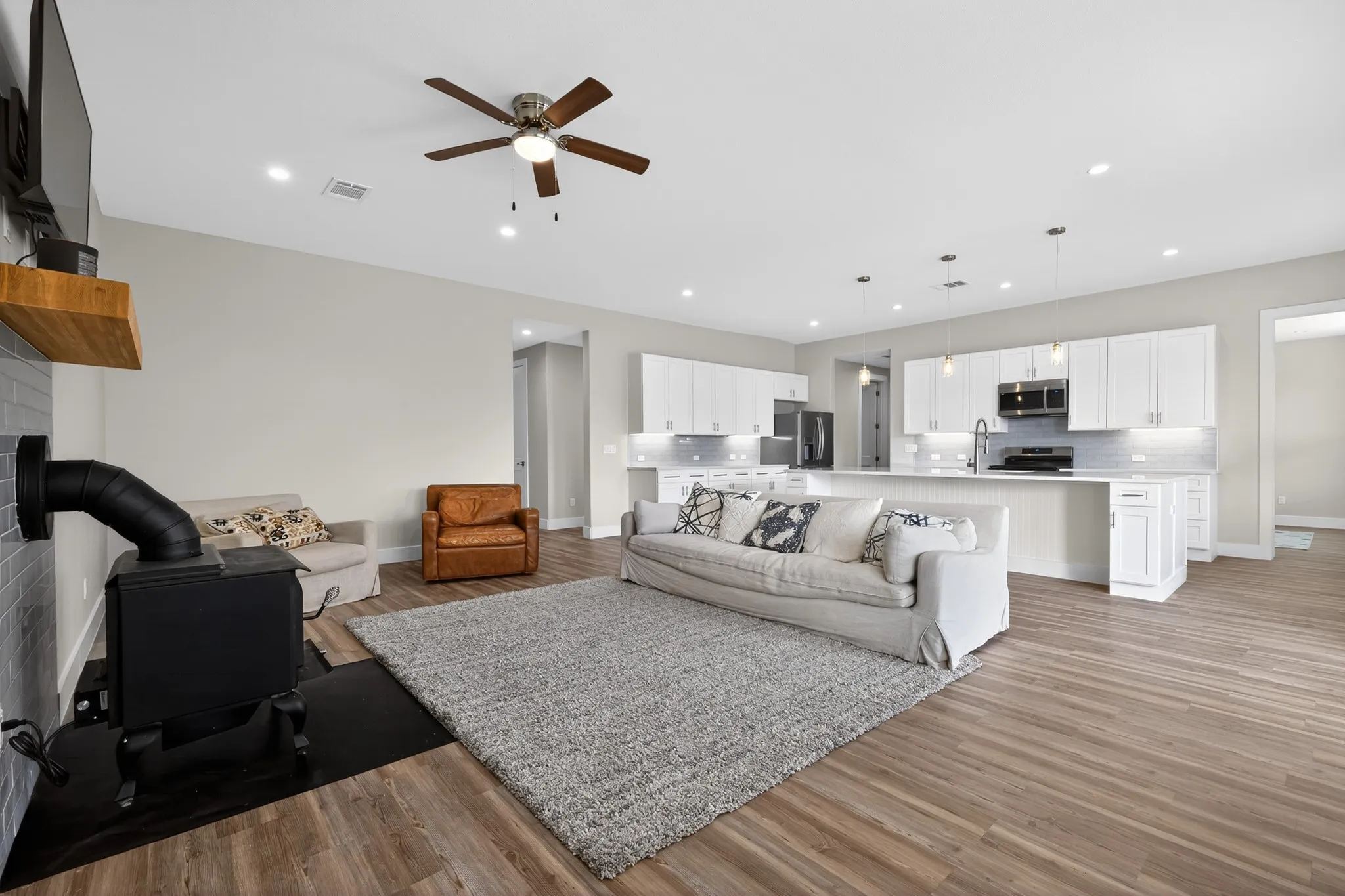 Open-concept design featuring wood-finish flooring, white cabinetry, a wood-burning stove, and a ceiling fan with integrated lighting