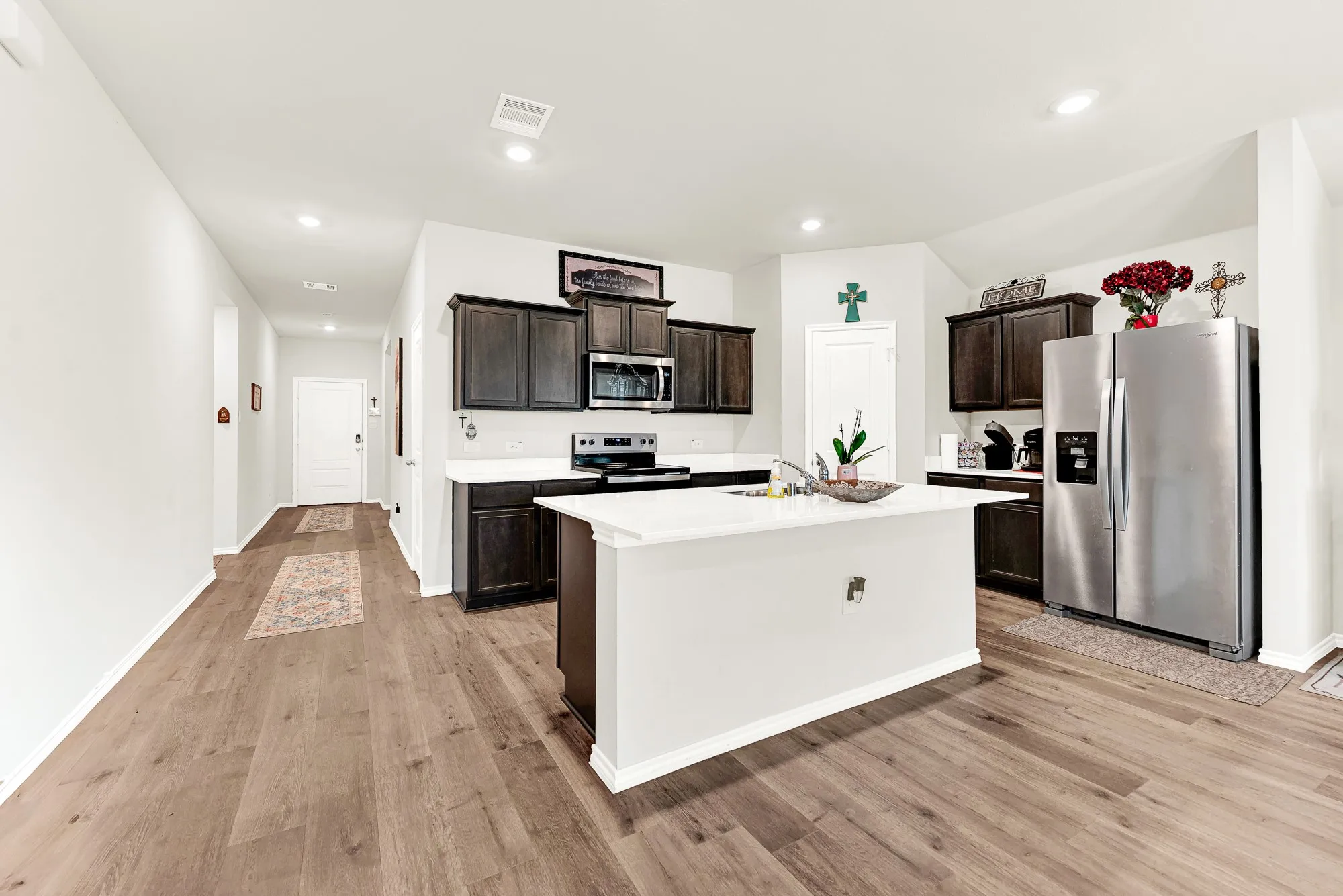 This light-filled kitchen features rich dark cabinetry, smooth white countertops, stainless steel appliances, and a central island set over wood-look flooring. It is an ideal setup for cooking weeknight meals while staying connected to family in nearby spaces.
