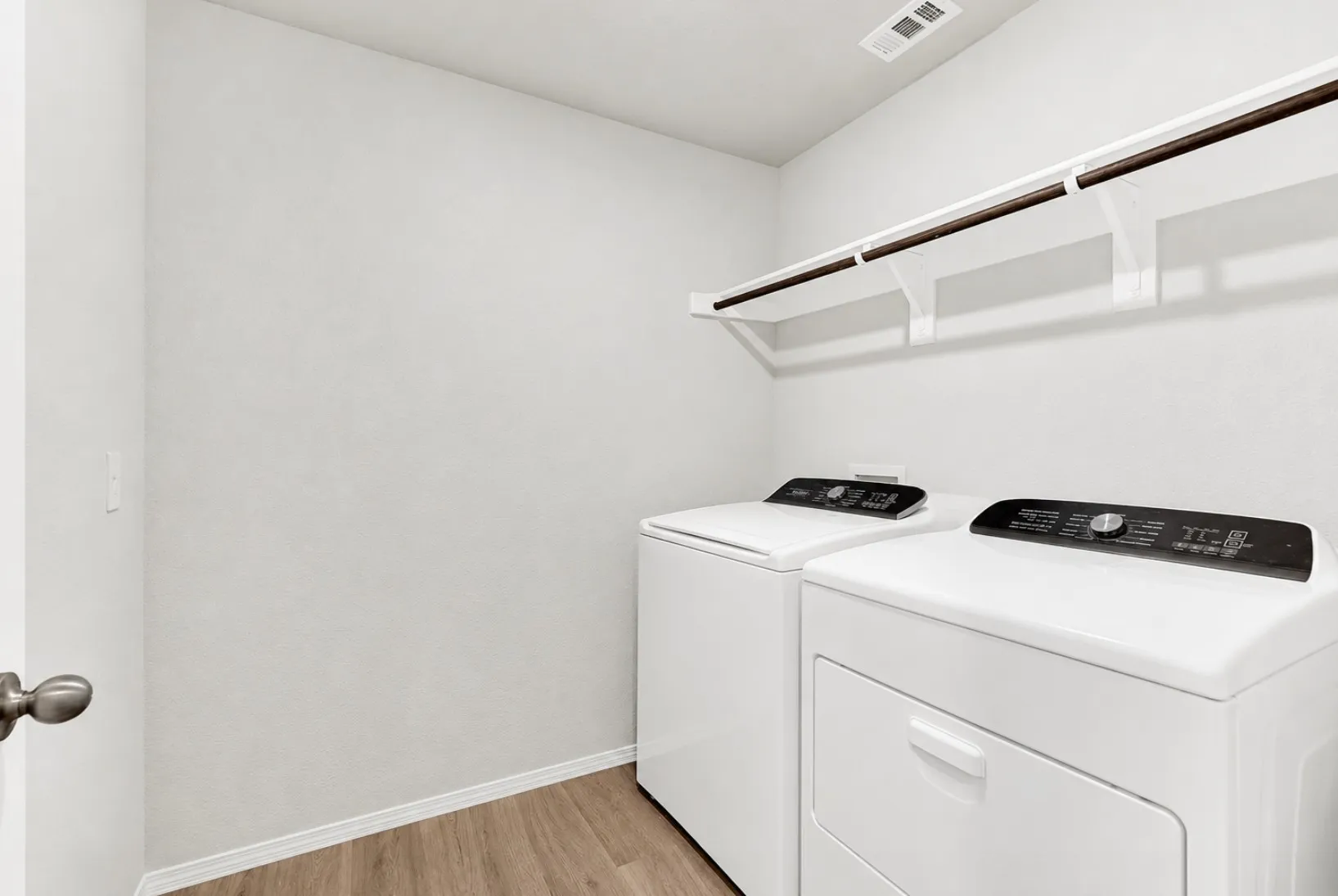This dedicated laundry room features durable wood-look flooring, full-size washer and dryer hookups, and a broad upper display shelf with an integrated hanging rod. It simplifies wash days with plenty of space to organize detergents and fold clothes.