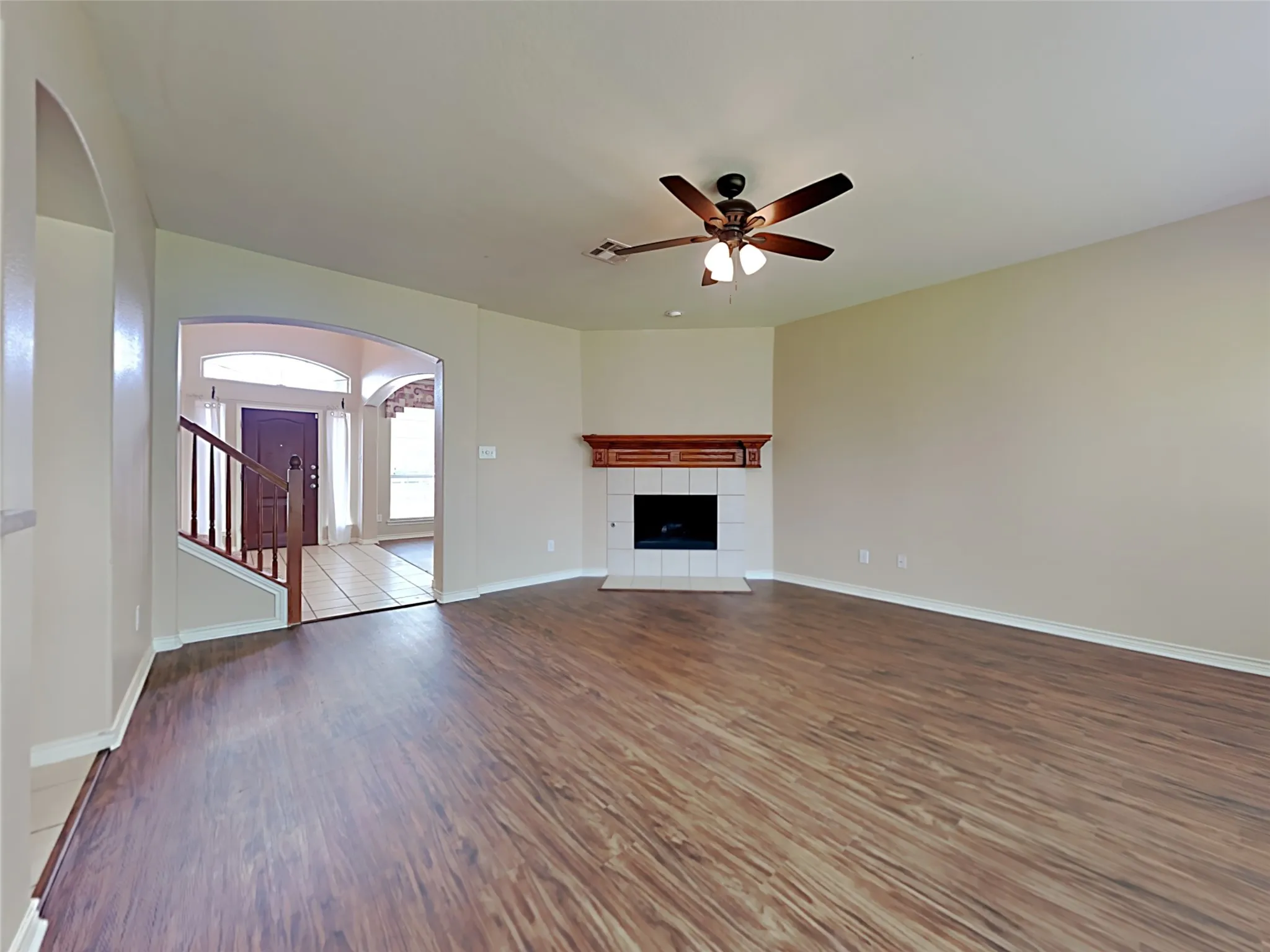 Wood-finish flooring, corner fireplace with tile surround and wood mantel, overhead ceiling fan with light, arched entryway, and staircase with wood railing