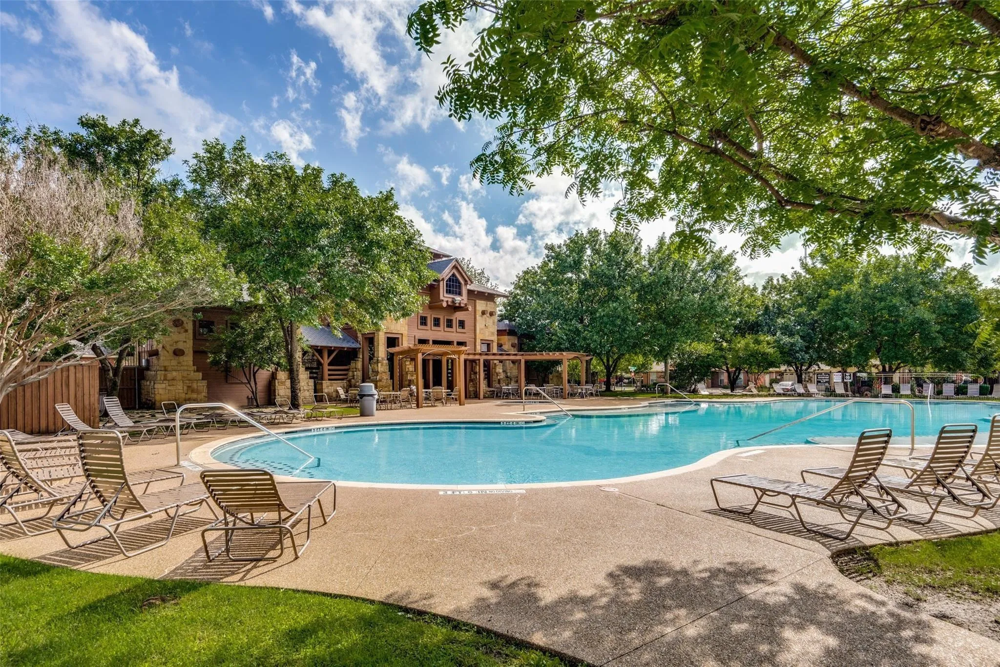 Expansive outdoor pool with a concrete surround, surrounded by mature trees and a stone-clad building
